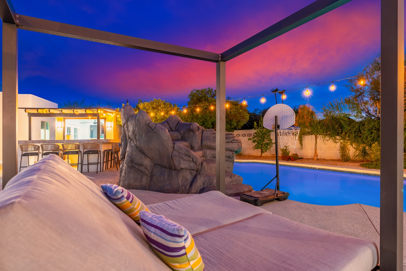 Unwind on your private poolside daybed as twilight paints the sky in vibrant hues, with sparkling lights and a basketball court completing your desert oasis retreat.
