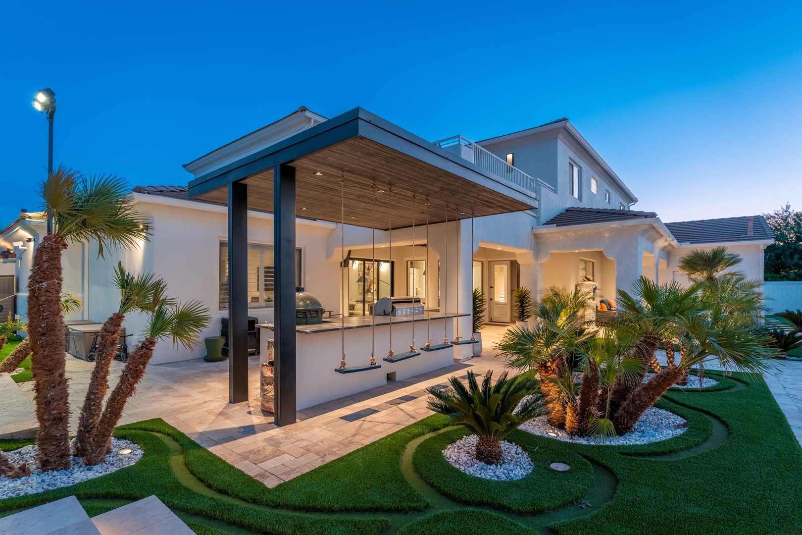 Modern vacation home at dusk, featuring a covered outdoor bar and lush tropical landscaping with illuminated palms and manicured lawns.