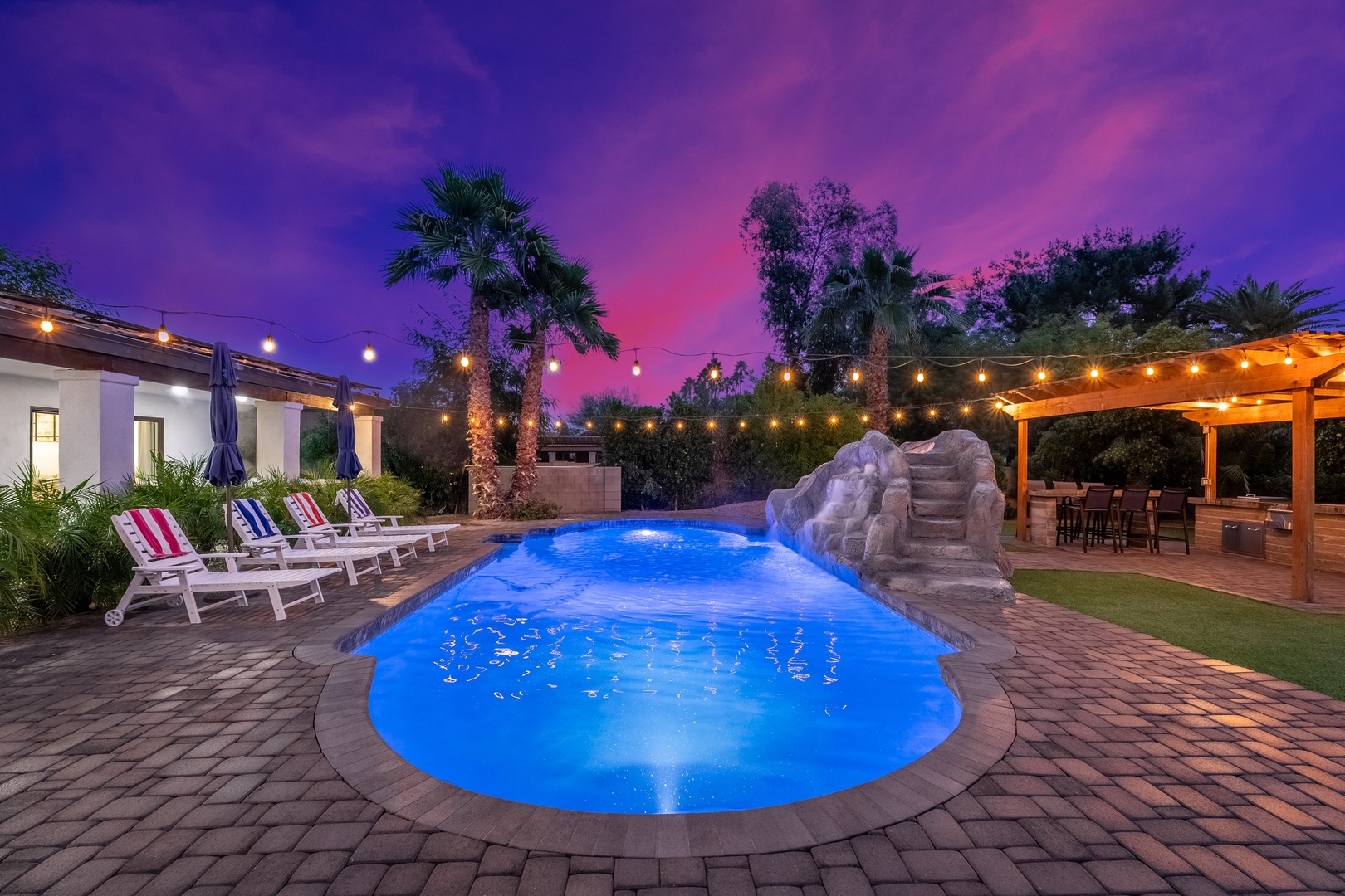Stunning twilight sets the scene for your desert oasis retreat. The glowing pool awaits your evening swims, while the covered patio and lush lawn provide perfect spaces to gather under string lights and vibrant skies.