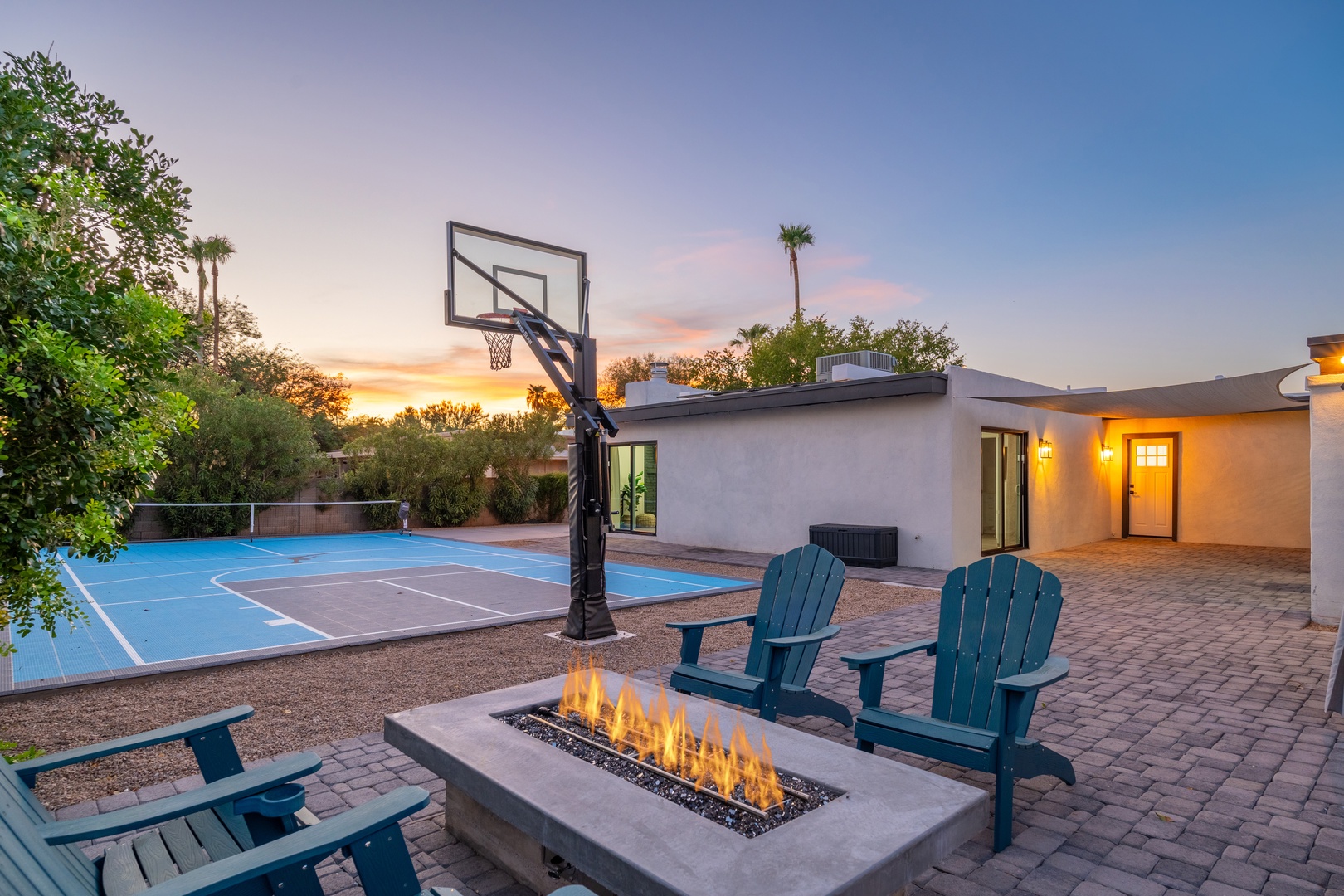 The property features a private outdoor recreation area with basketball court, outdoor fire pit, and contemporary seating, set against a scenic backdrop during golden hour.