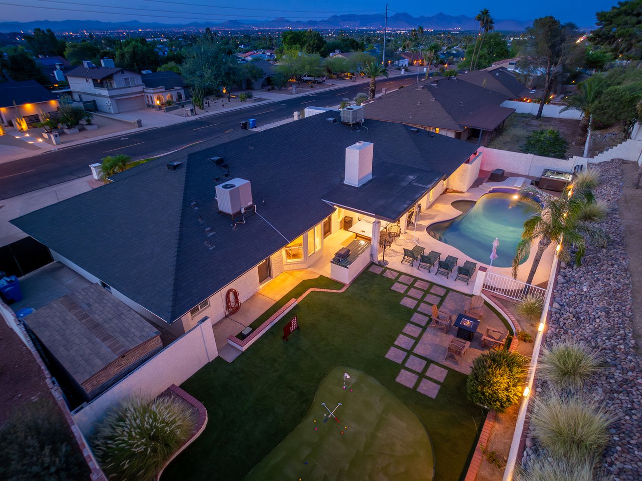Aerial view of a vacation rental property showcasing a spacious backyard with pool, putting green, and multiple outdoor entertaining areas set against mountain views at twilight.