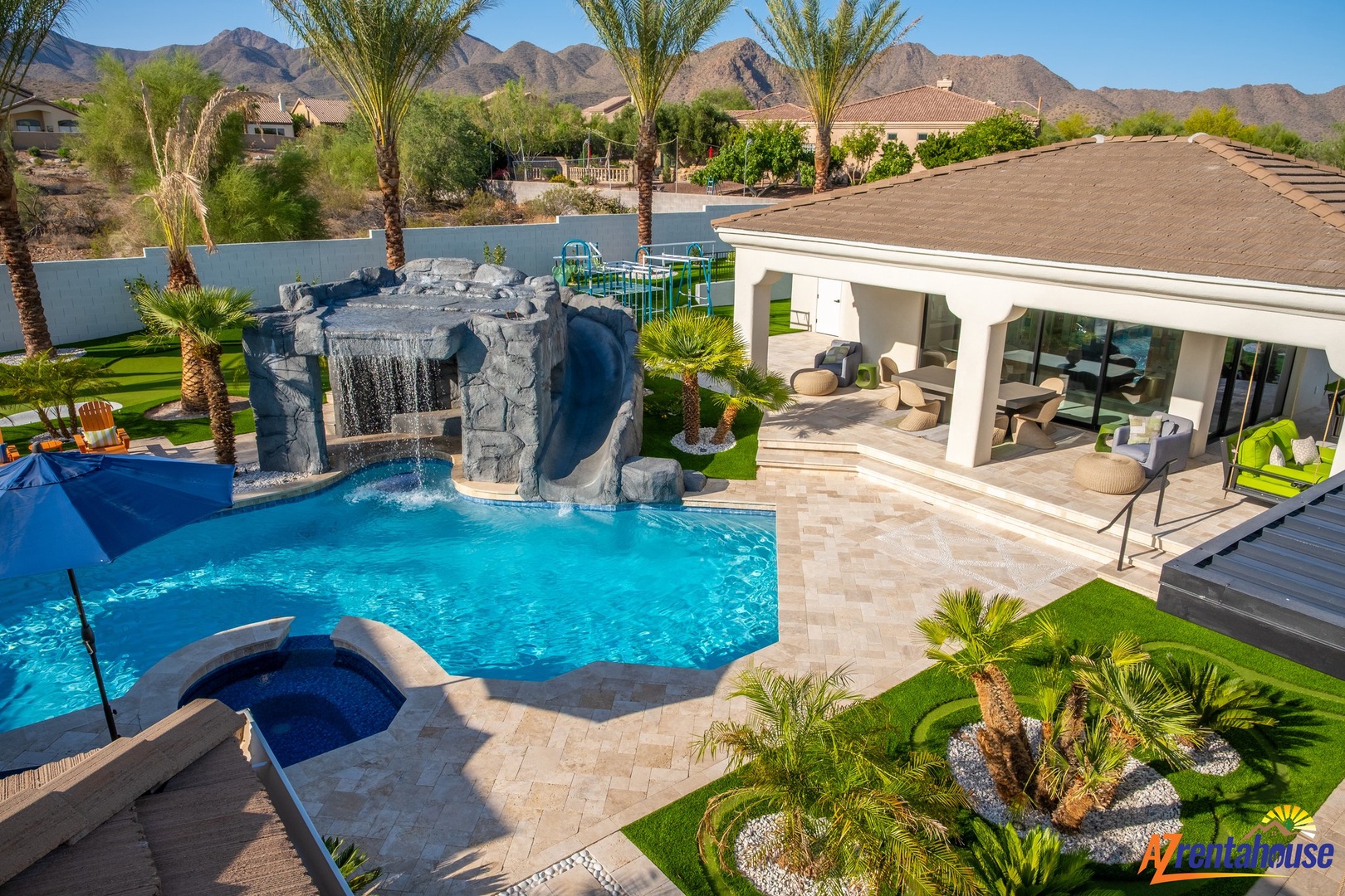 Luxurious desert oasis with dramatic mountain backdrop featuring resort-style pool, waterfall grotto, spa, covered patio, and manicured tropical landscaping.