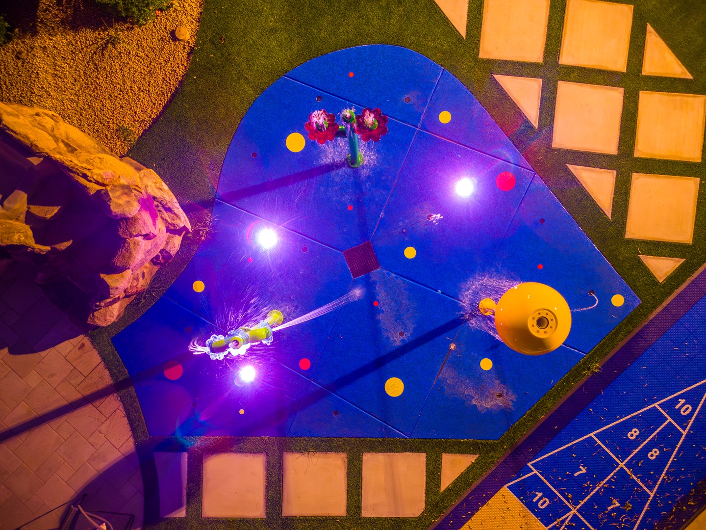 An aerial view at night reveals a vibrant illuminated swimming pool with playful water features and colorful floating elements, creating a festive resort atmosphere.