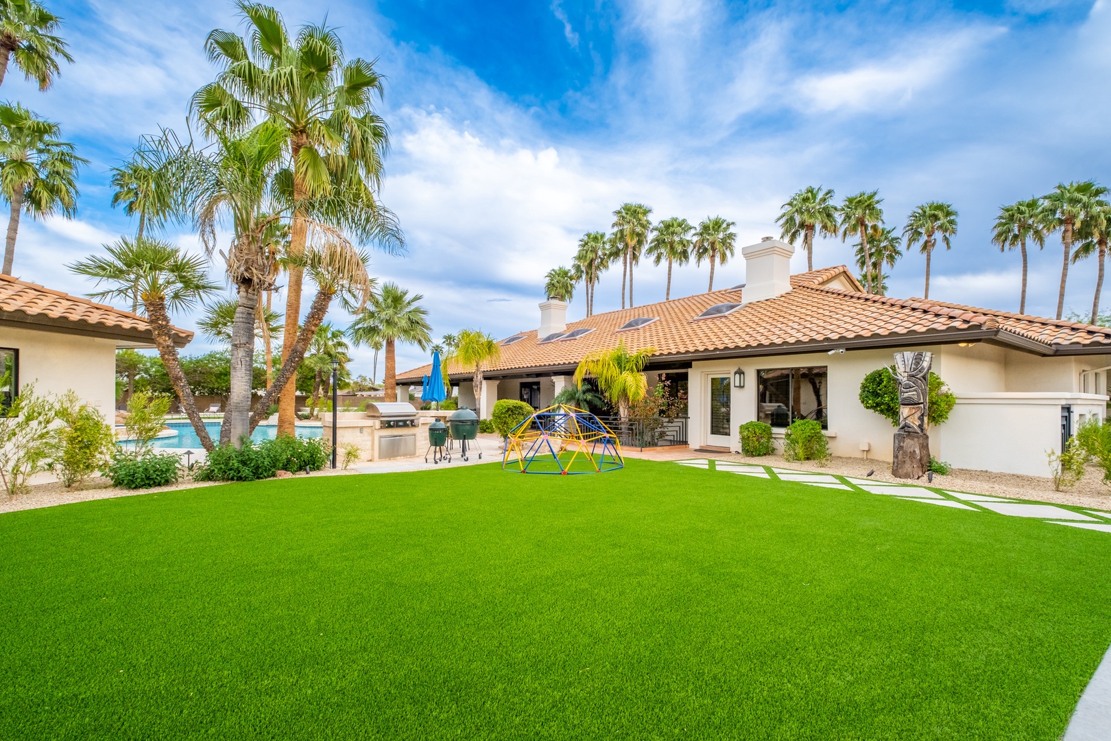 A sun-drenched desert oasis features Mediterranean-style architecture surrounded by lush palms and verdant lawn, with a sparkling pool area perfect for outdoor enjoyment.