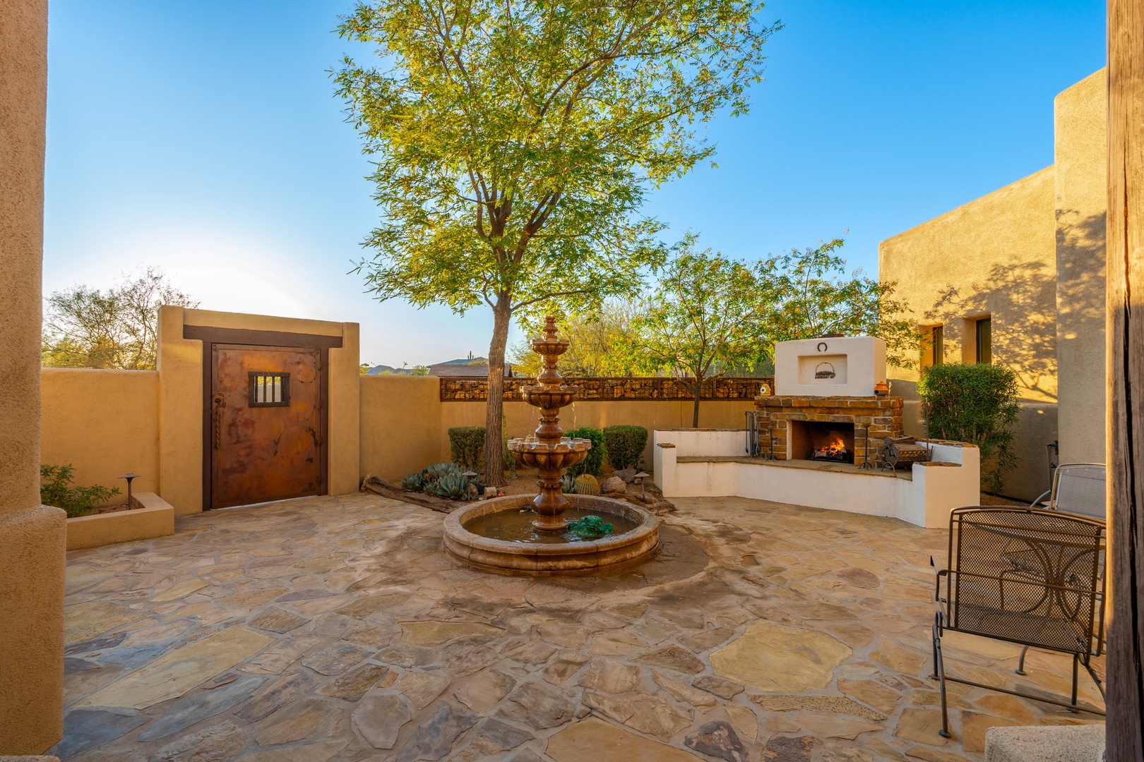 An inviting courtyard oasis awaits with a soothing fountain centerpiece, shaded by a graceful tree, and an outdoor fireplace for serene desert evenings under open skies.