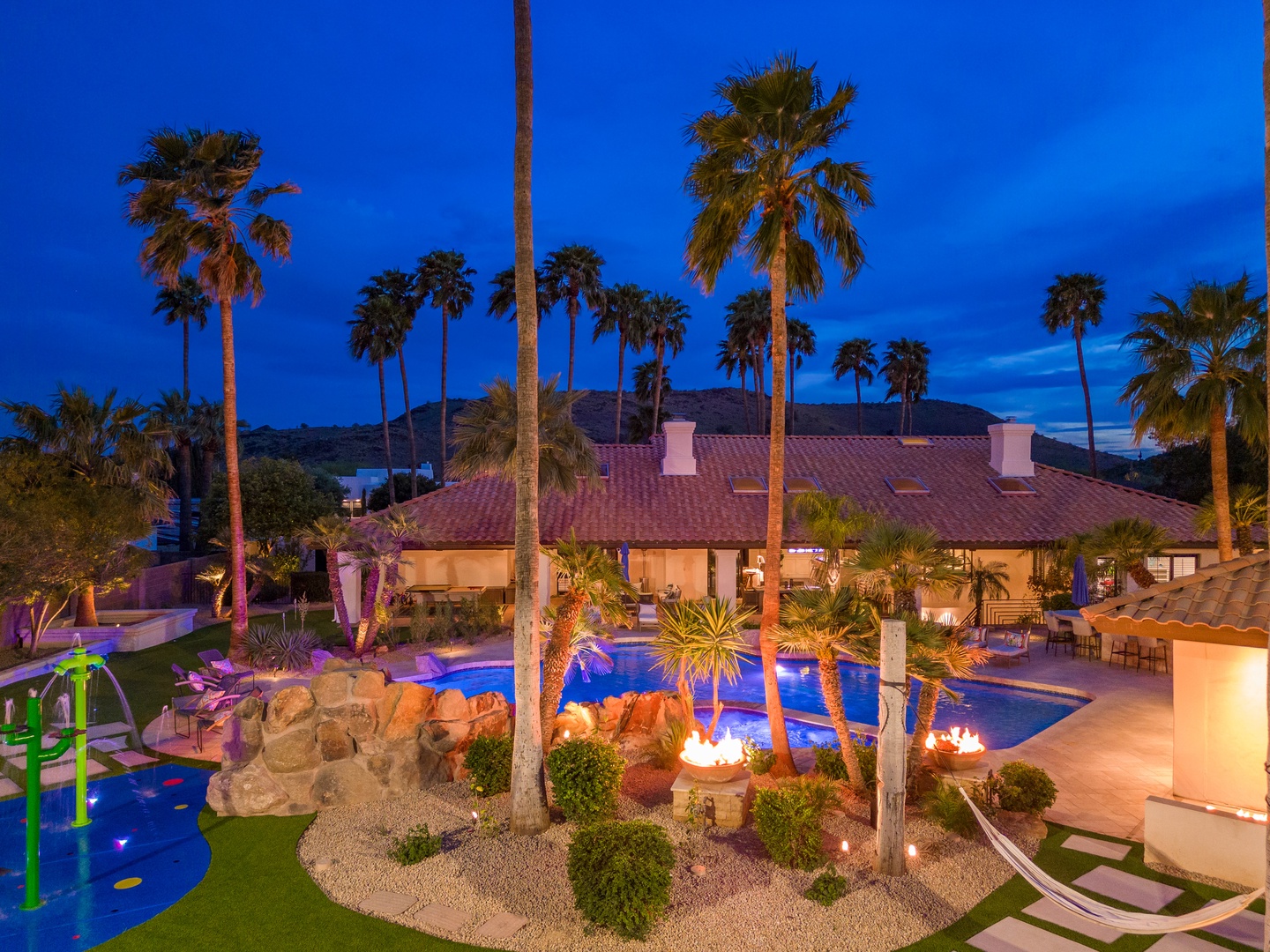 An enchanting twilight view showcases the property's stunning backyard oasis, featuring a sparkling pool, tropical palms, outdoor lounge spaces, and fire features beneath a vivid blue desert sky.