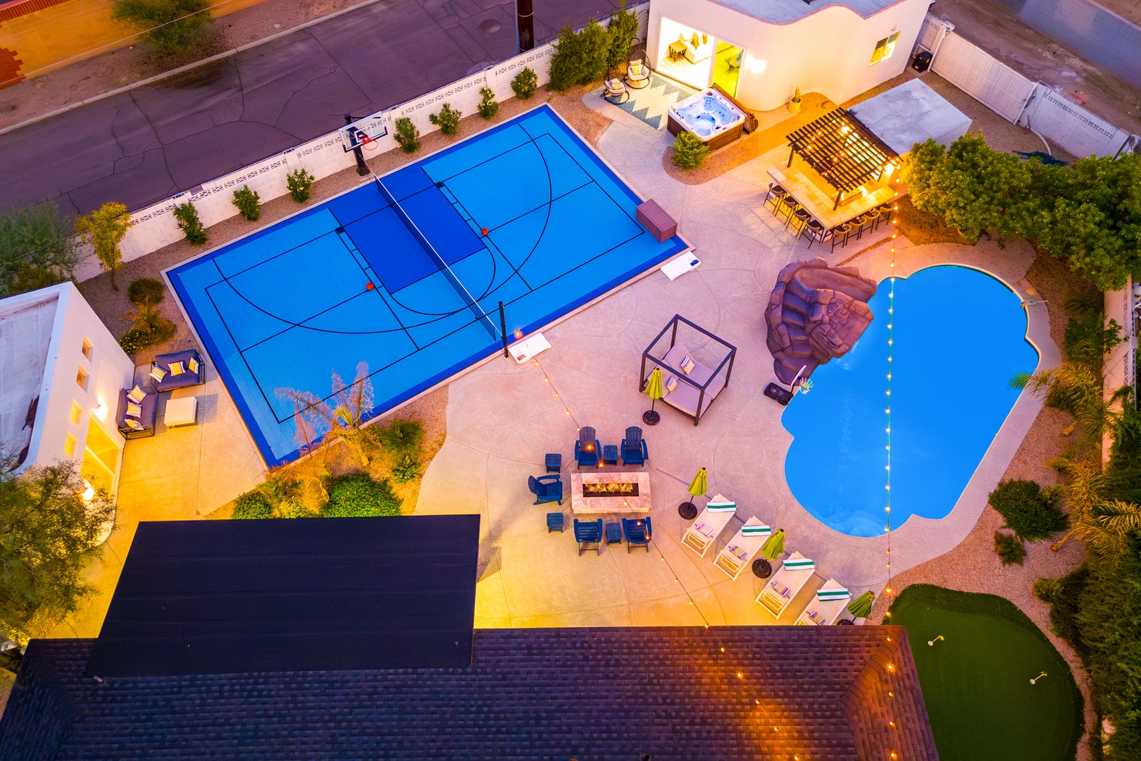 An aerial view showcases this desert resort's impressive recreation area featuring a basketball court, free-form swimming pool, hot tub, and outdoor dining pavilion at dusk.