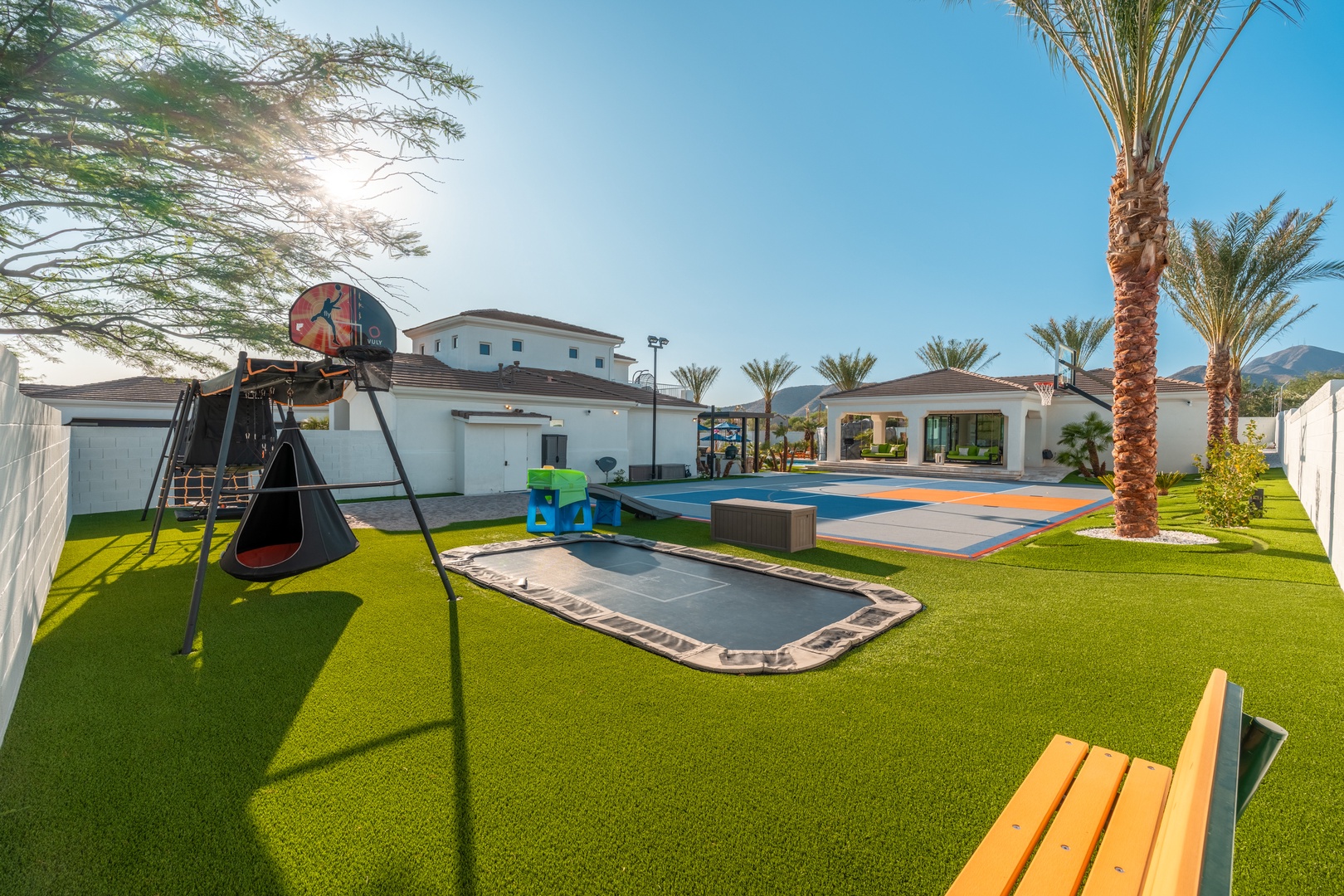 Expansive backyard resort designed for families, featuring a sparkling pool with sport court, in-ground trampoline, basketball hoop, swings, and lush synthetic grass under towering palms.