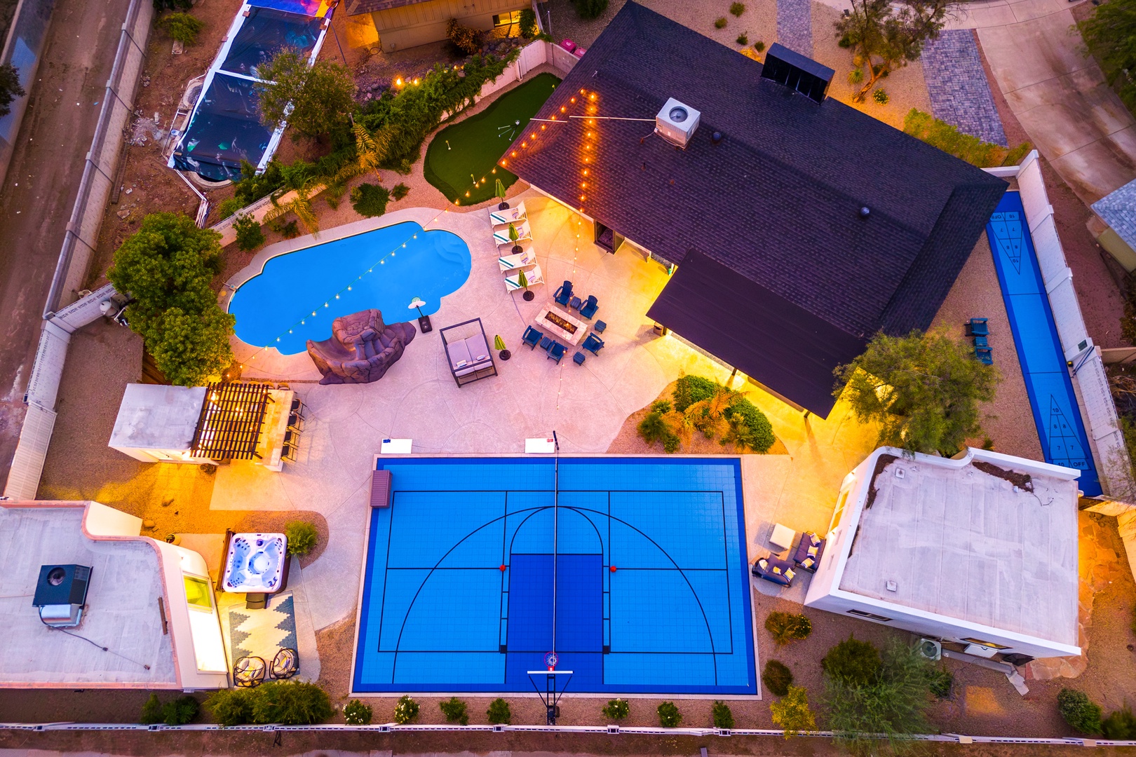 Aerial view showcasing a vacation rental property with diverse outdoor amenities including a swimming pool, sport court, hot tub, putting green, and covered patio area surrounded by mature landscaping and neighboring residences.