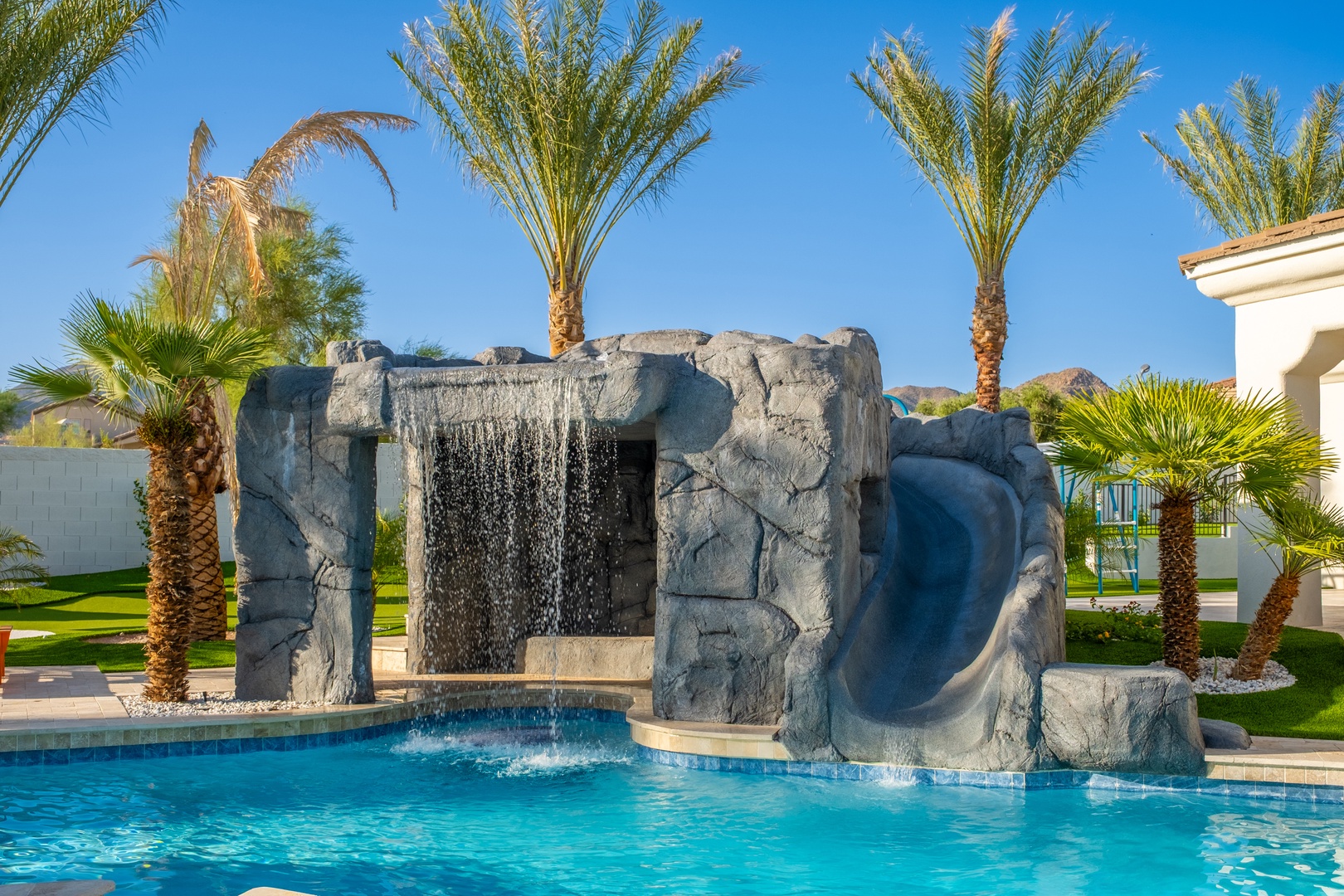 A sparkling pool with a dramatic rock waterfall feature, framed by swaying palms and desert mountains. This stunning outdoor oasis offers a serene retreat in the sunshine.