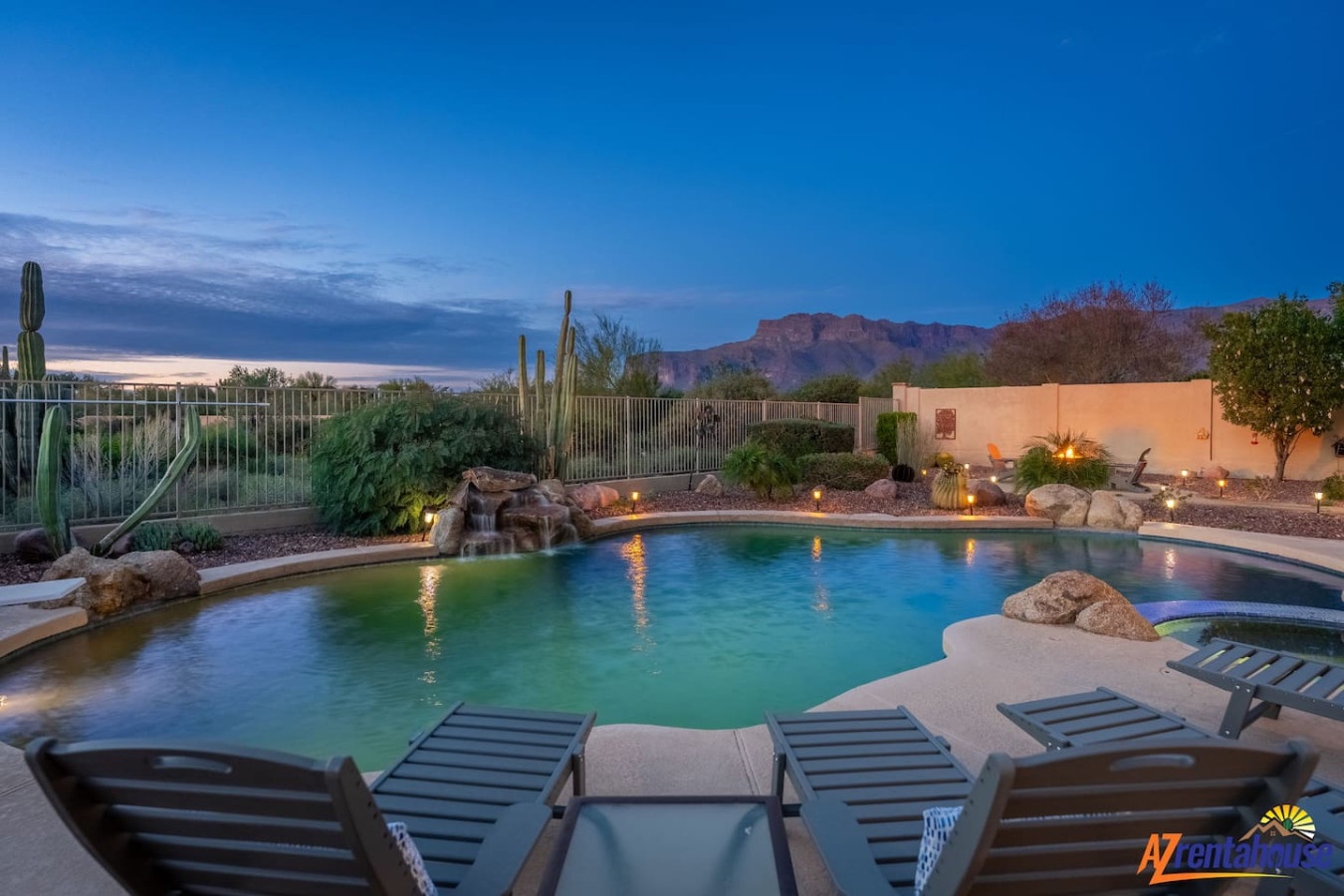 Desert oasis with stunning mountain backdrop features an illuminated pool and dramatic red rock formations on the horizon.