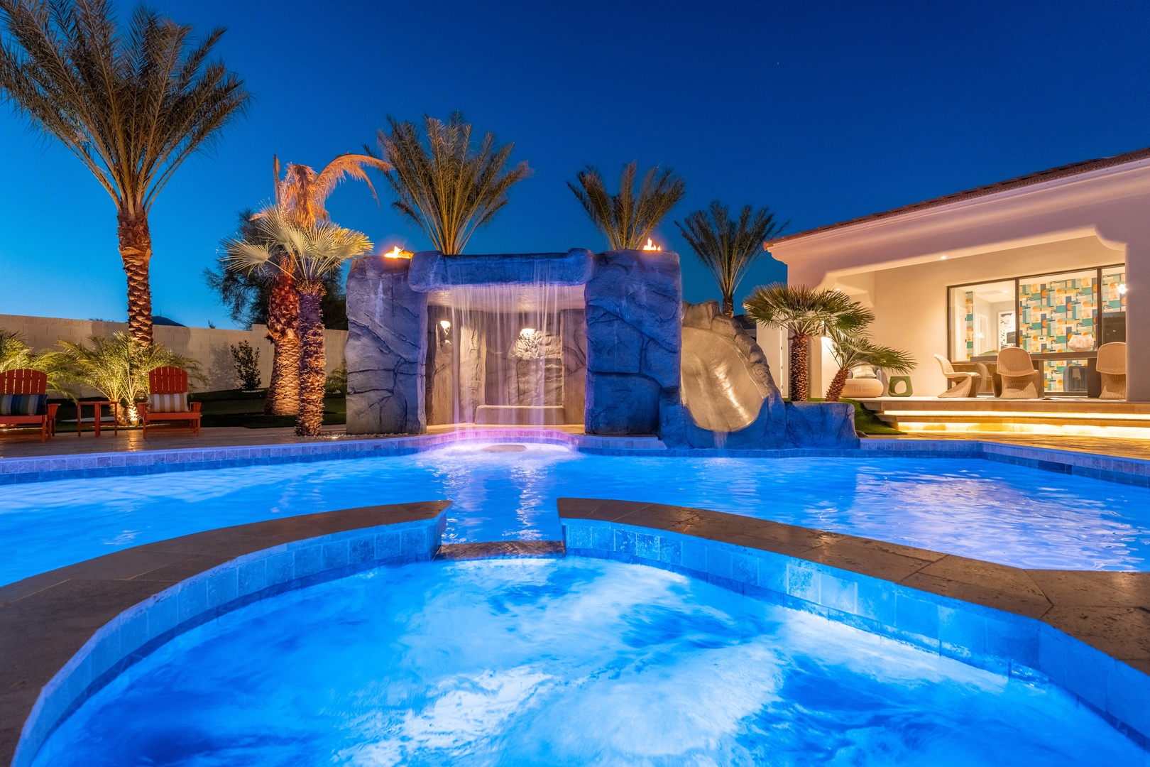 An illuminated tropical oasis at dusk featuring an elegant waterfall fountain, glowing pool, hot tub, and palm trees under evening skies, with modern entertaining spaces nearby.