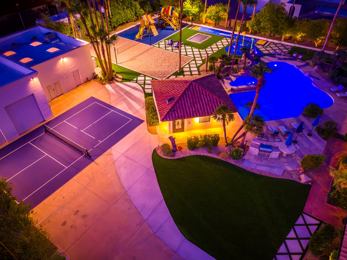 Aerial view of the vacation property at night, showcasing a resort-style backyard with swimming pool, tennis court, playground, and lush landscaping—a private entertainment paradise.