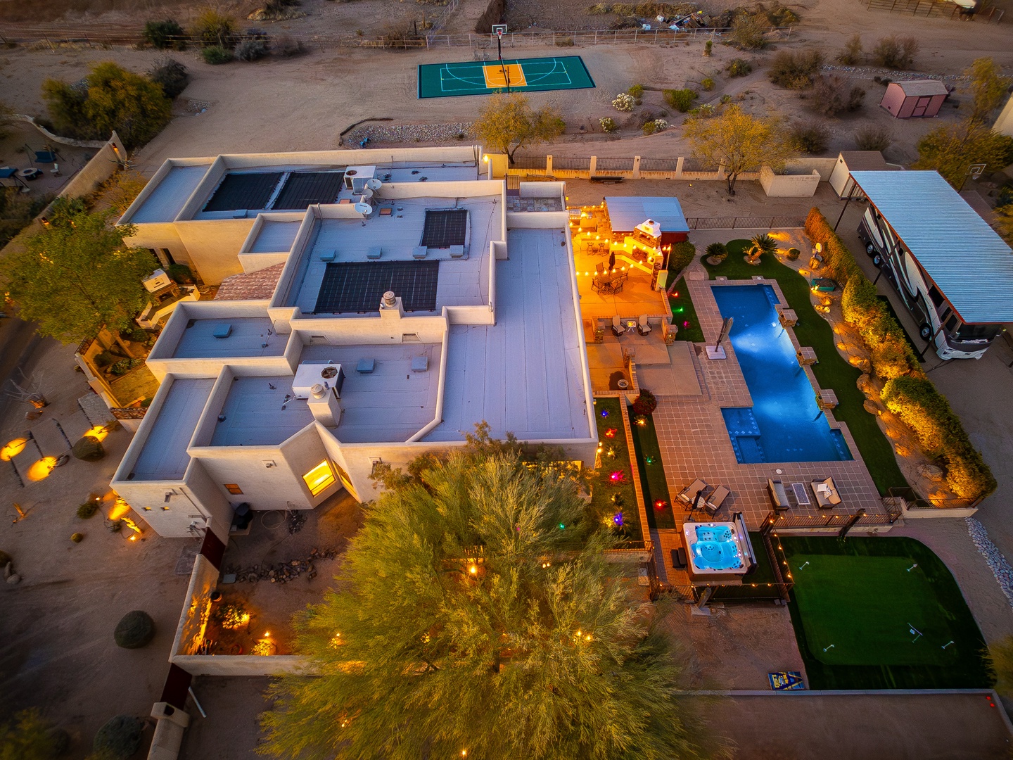 Aerial view of a modern desert vacation rental featuring a lap pool, hot tub, outdoor dining areas, basketball court, and sprawling grounds illuminated at dusk.