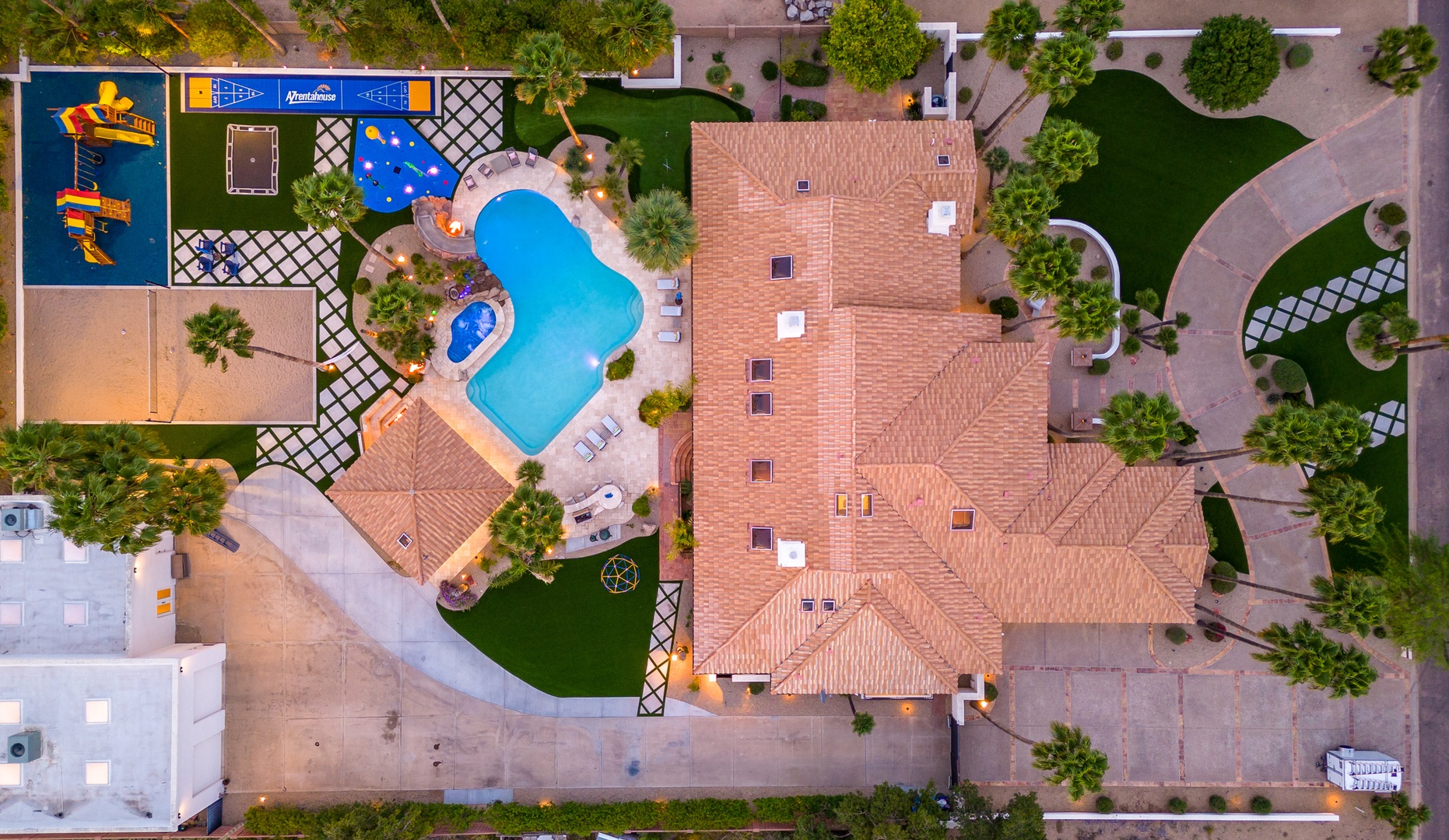 Aerial view showcasing the property's layout with a freeform pool, children's play area with colorful water slides, manicured lawns, palm trees, and tile-roofed architecture within a landscaped residential setting.