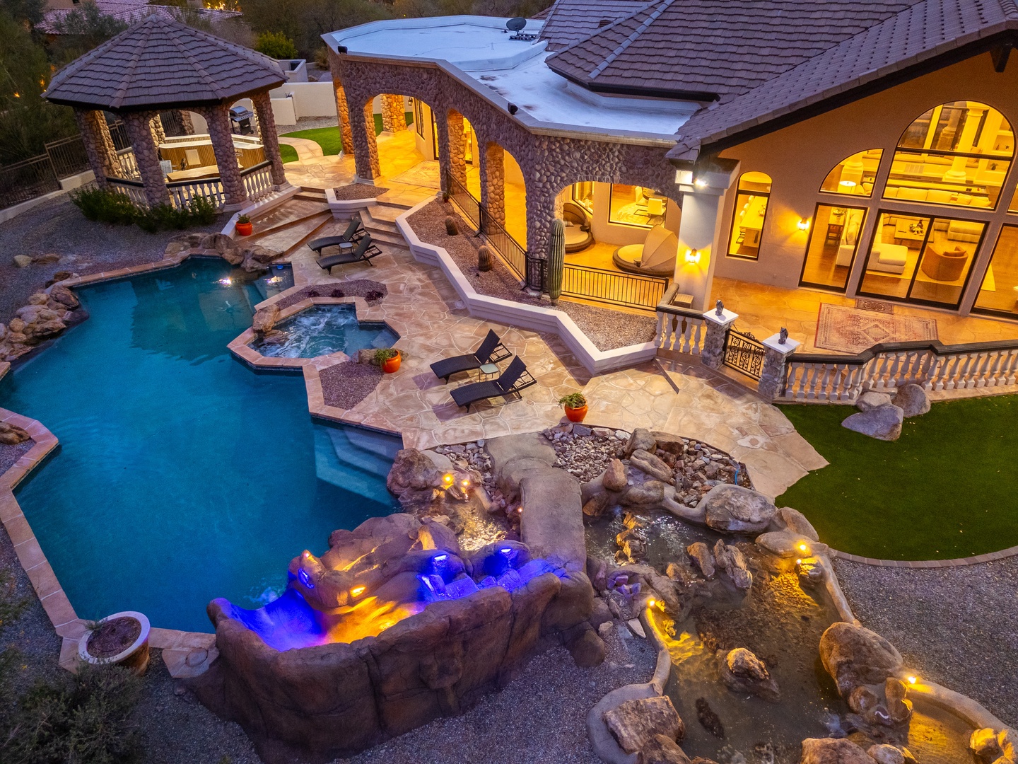 An aerial view captures the property's luxurious exterior at dusk, featuring a resort-style pool, spa with illuminated water features, covered gazebo, and spacious stone patio set against the hillside.