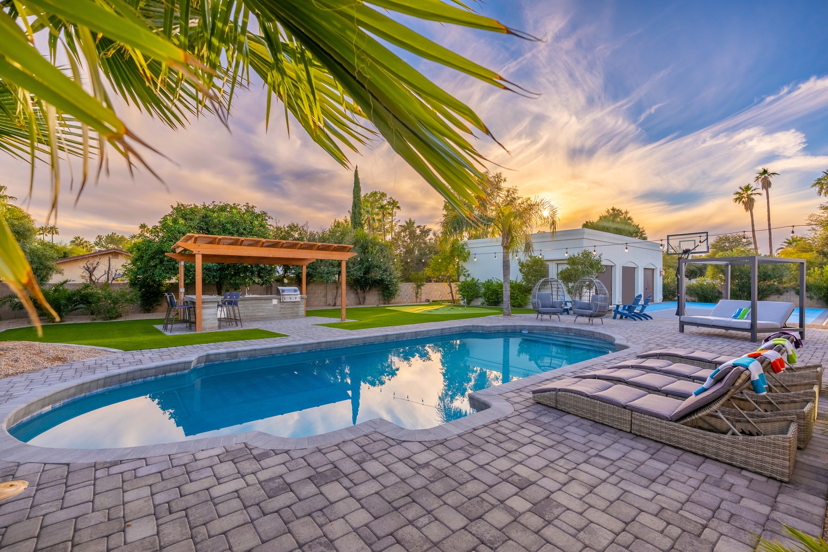 A private backyard oasis featuring a sparkling pool framed by palm trees, pergola-covered dining, and plush lounge beds under golden sunset skies.