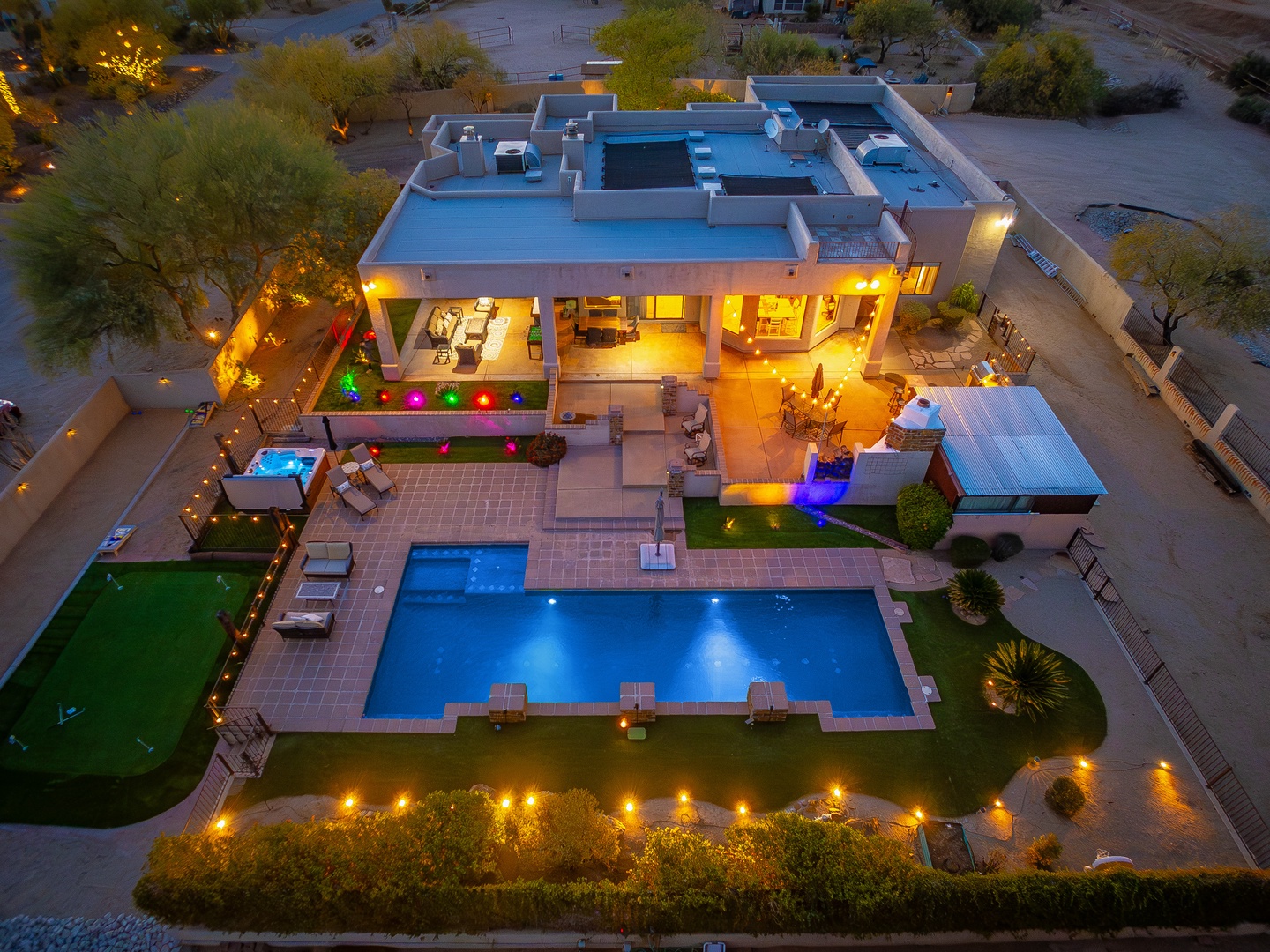 A stunning desert vacation home features modern architecture with expansive patios, a sparkling swimming pool, colorful lighting, and scenic desert landscaping at twilight.