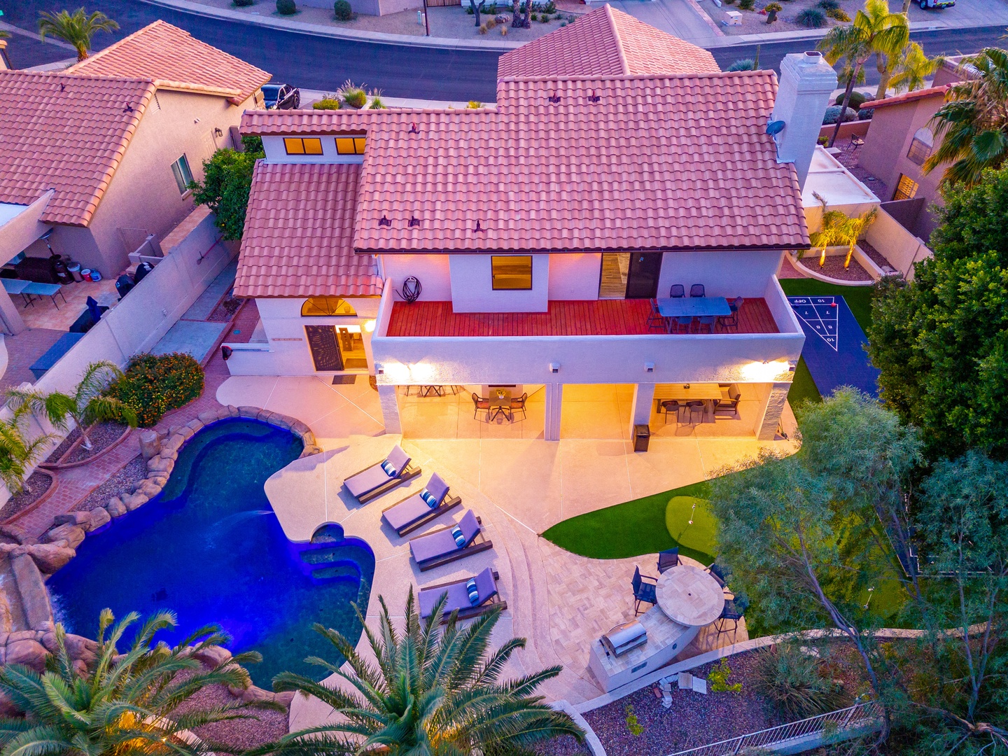 Aerial view of a vacation rental property featuring a lagoon-style pool, covered patio with dining areas, and Spanish tile roof surrounded by palm trees and desert landscaping.