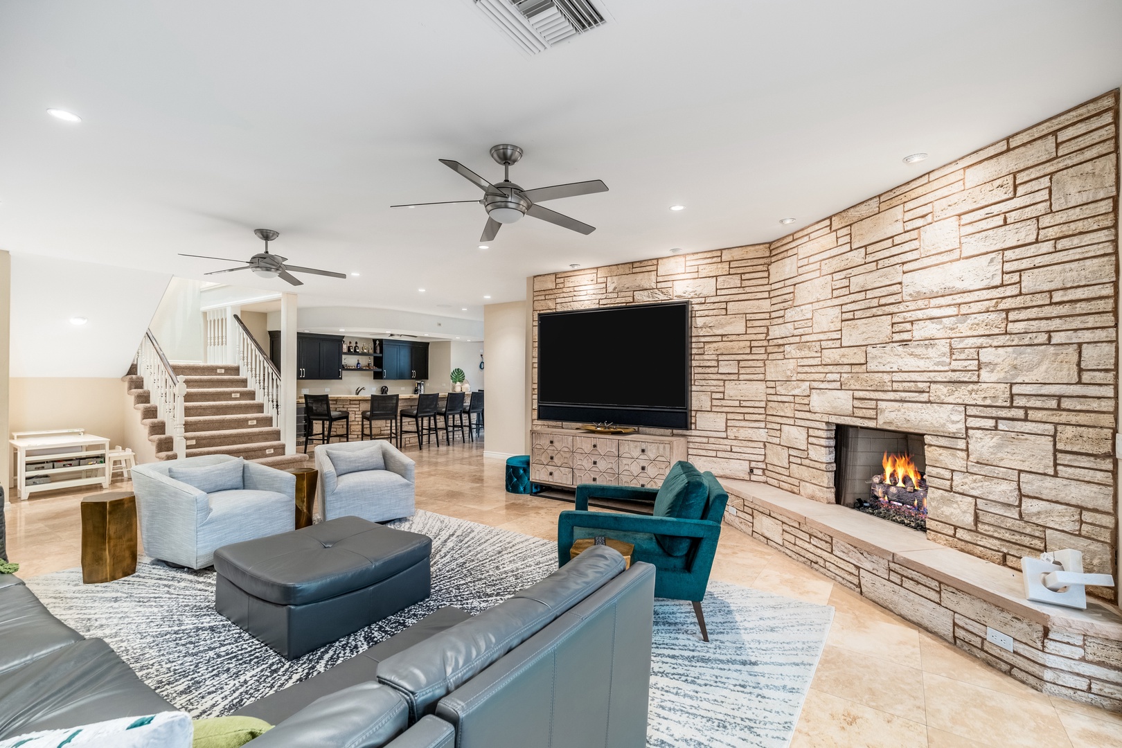 Relax in your expansive living room featuring a stunning stone fireplace, modern furnishings, and open-concept design connecting to the kitchen—perfect for unwinding or entertaining guests.
