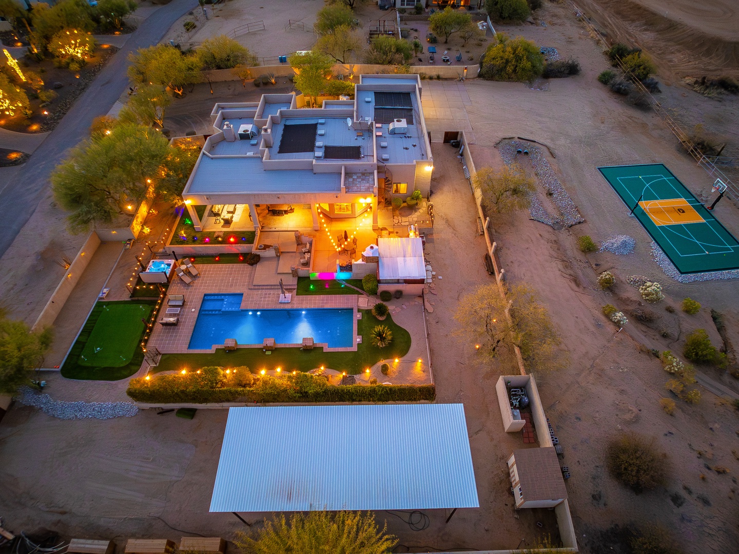 Aerial view of the property at dusk, showcasing the modern desert retreat with illuminated pool, tennis court, outdoor seating areas, and surrounded by natural landscape.