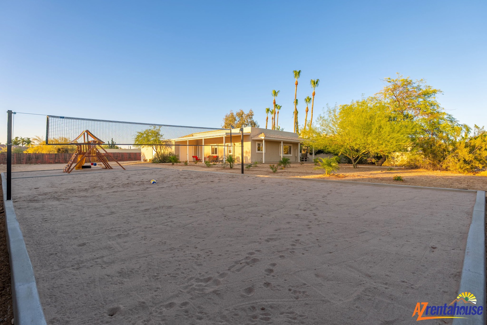 A spacious sand volleyball court with a playground creates an inviting outdoor recreation area, set against a backdrop of mature palm trees and desert landscaping.