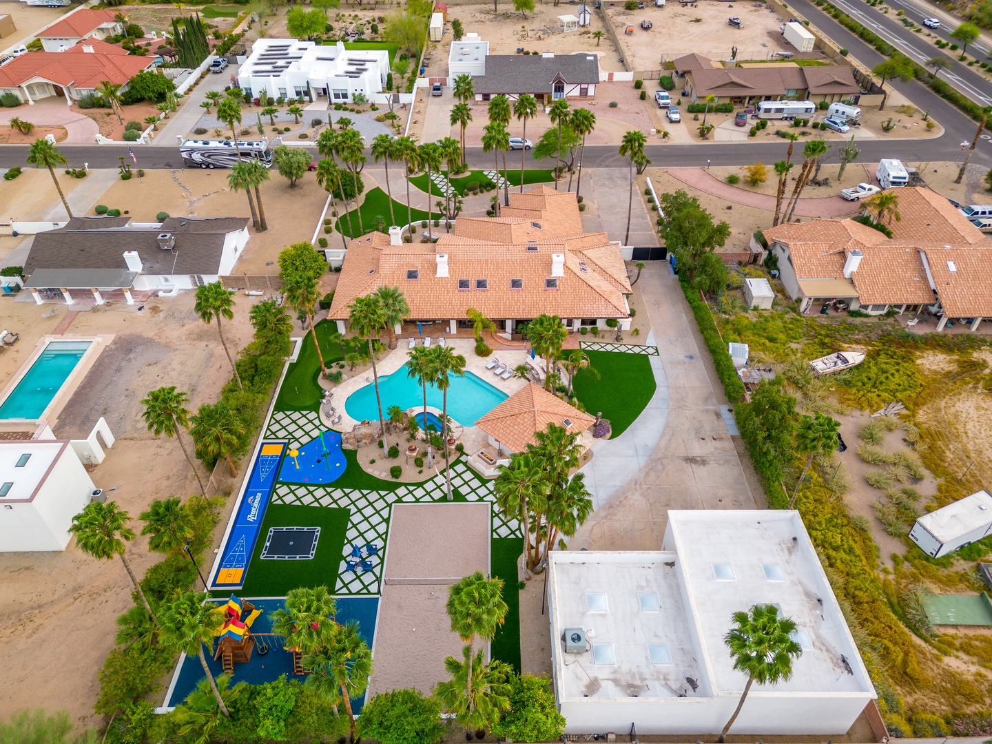 Aerial view of a spacious vacation property featuring a sparkling pool, colorful playground, lush palm trees, and charming Spanish-style architecture in a quiet residential neighborhood.