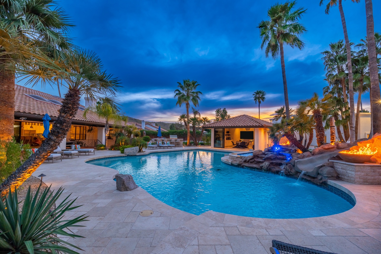 Resort-style pool with dramatic twilight skies and swaying palms, surrounded by warmly lit buildings and a cozy fire feature.