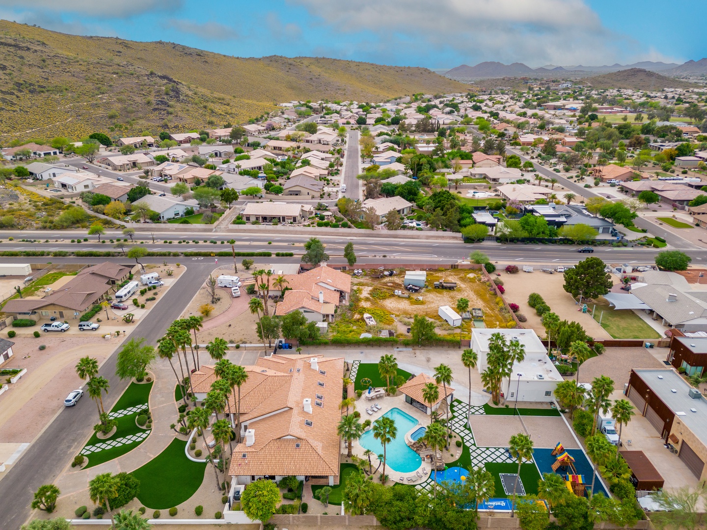 Aerial view of the property nestled in a residential neighborhood, surrounded by desert hills and mountain vistas. The pool and lush palm trees stand out in this sunny Arizona setting.