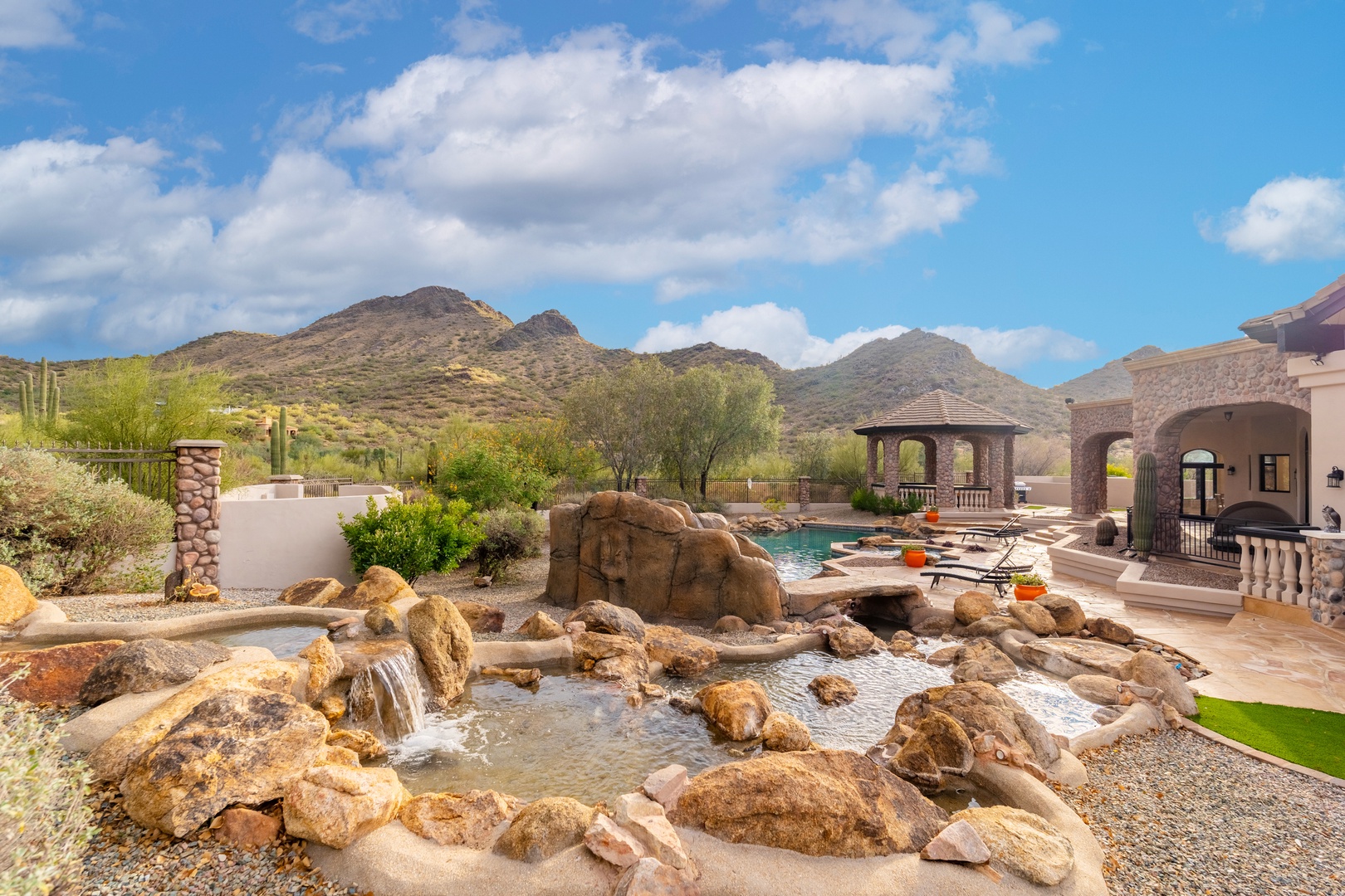 Dramatic mountain views frame a desert oasis featuring a natural stone waterfall, tranquil pool, and shaded ramada—your private sanctuary under Arizona skies.