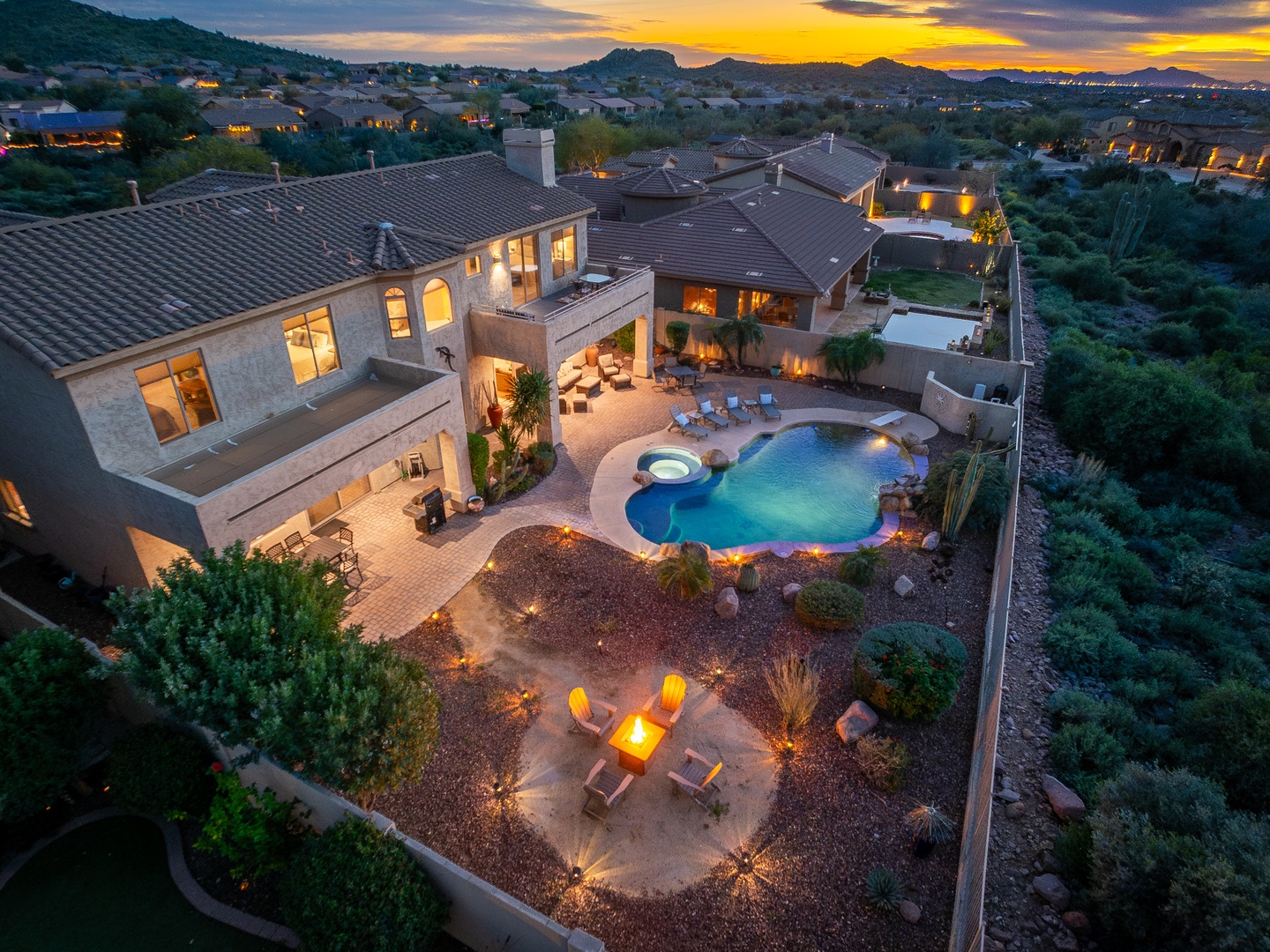 Stunning desert estate at twilight featuring a resort-style pool, hot tub, outdoor fire pit, and multiple lounging areas, all framed by mountain vistas and breathtaking sunset views.
