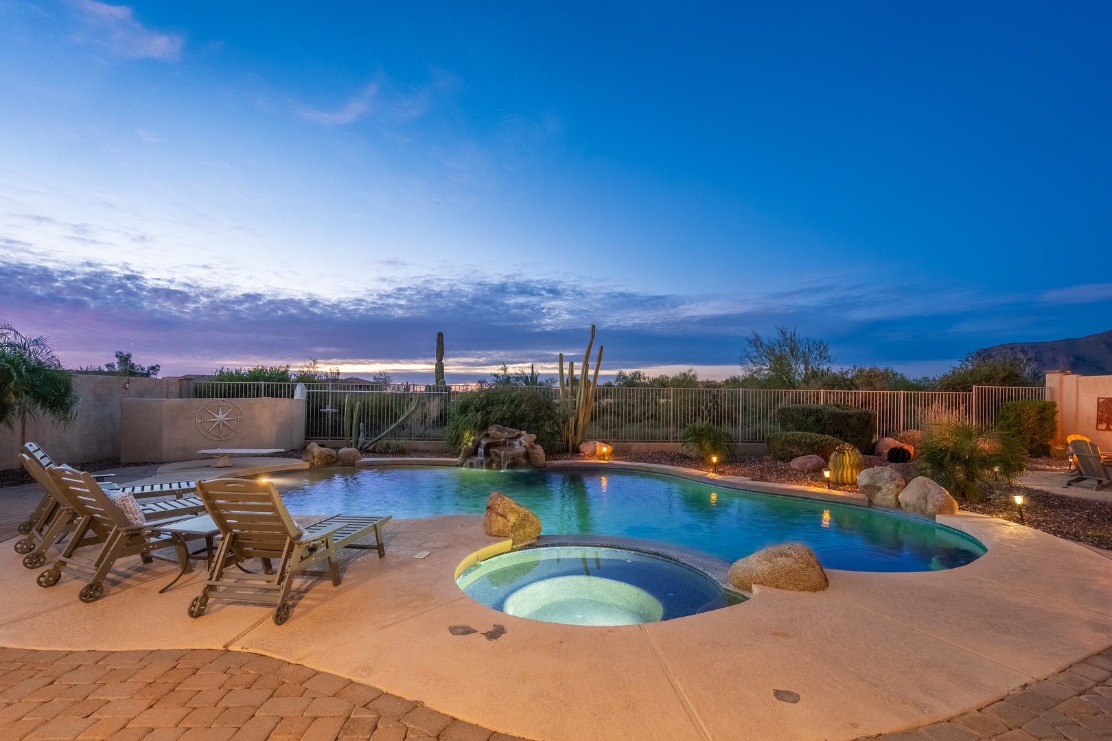 A stunning desert oasis awaits with a heated pool, soothing hot tub, and glowing landscape lighting set against dramatic twilight skies and mountain views.