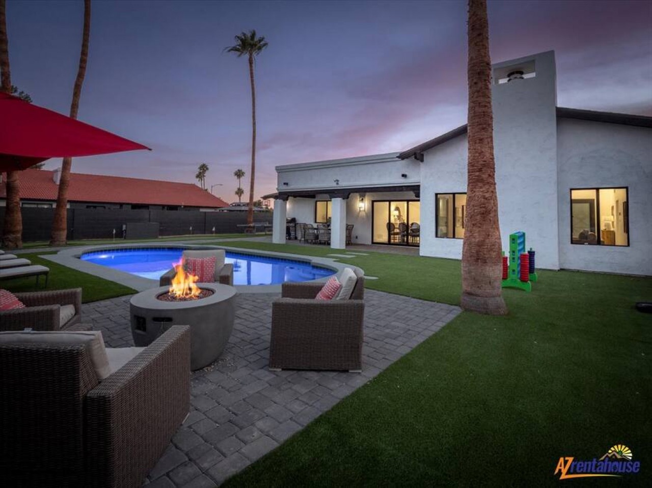 Modern desert oasis featuring a sparkling pool, fire pit seating area, and contemporary architecture under purple-hued twilight skies.