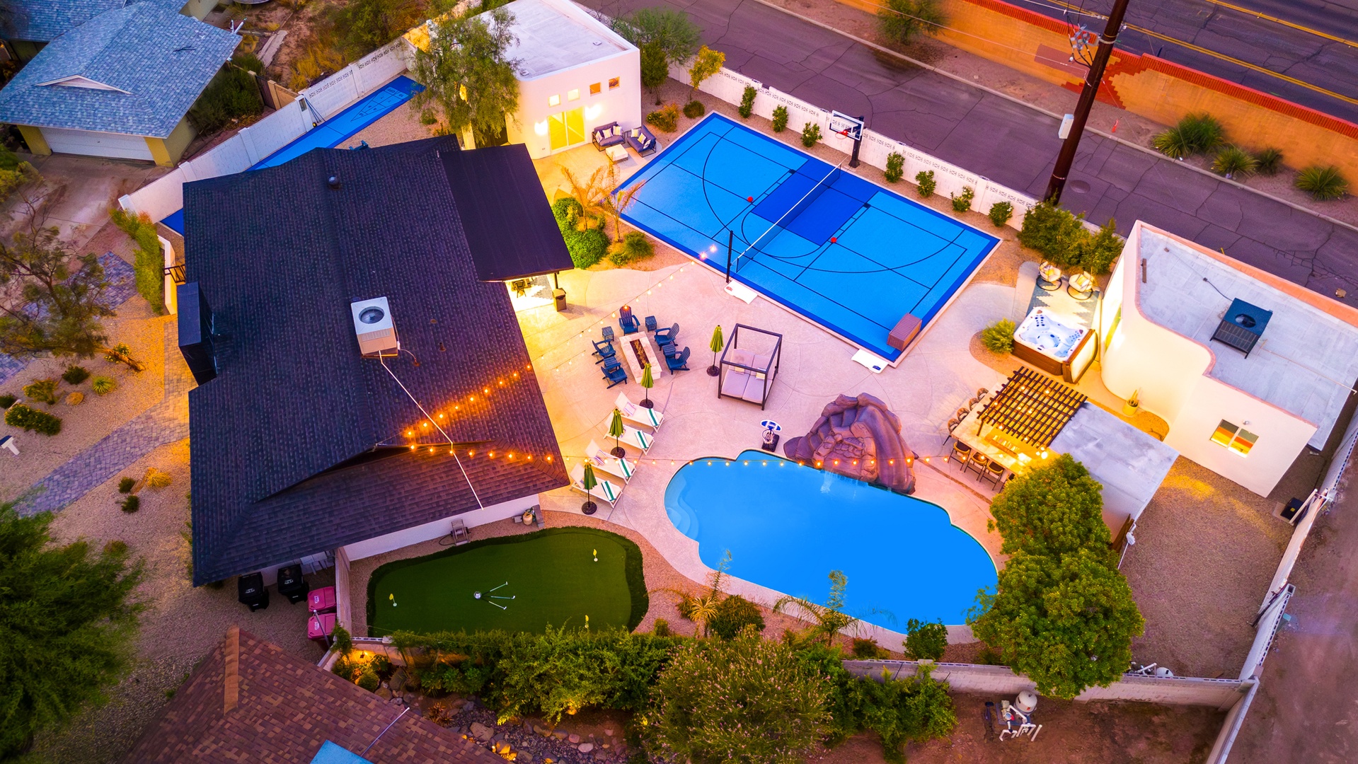 Aerial view of the property showcasing an expansive outdoor entertainment area with tennis court, swimming pool, putting green, and landscaped patio spaces at twilight.