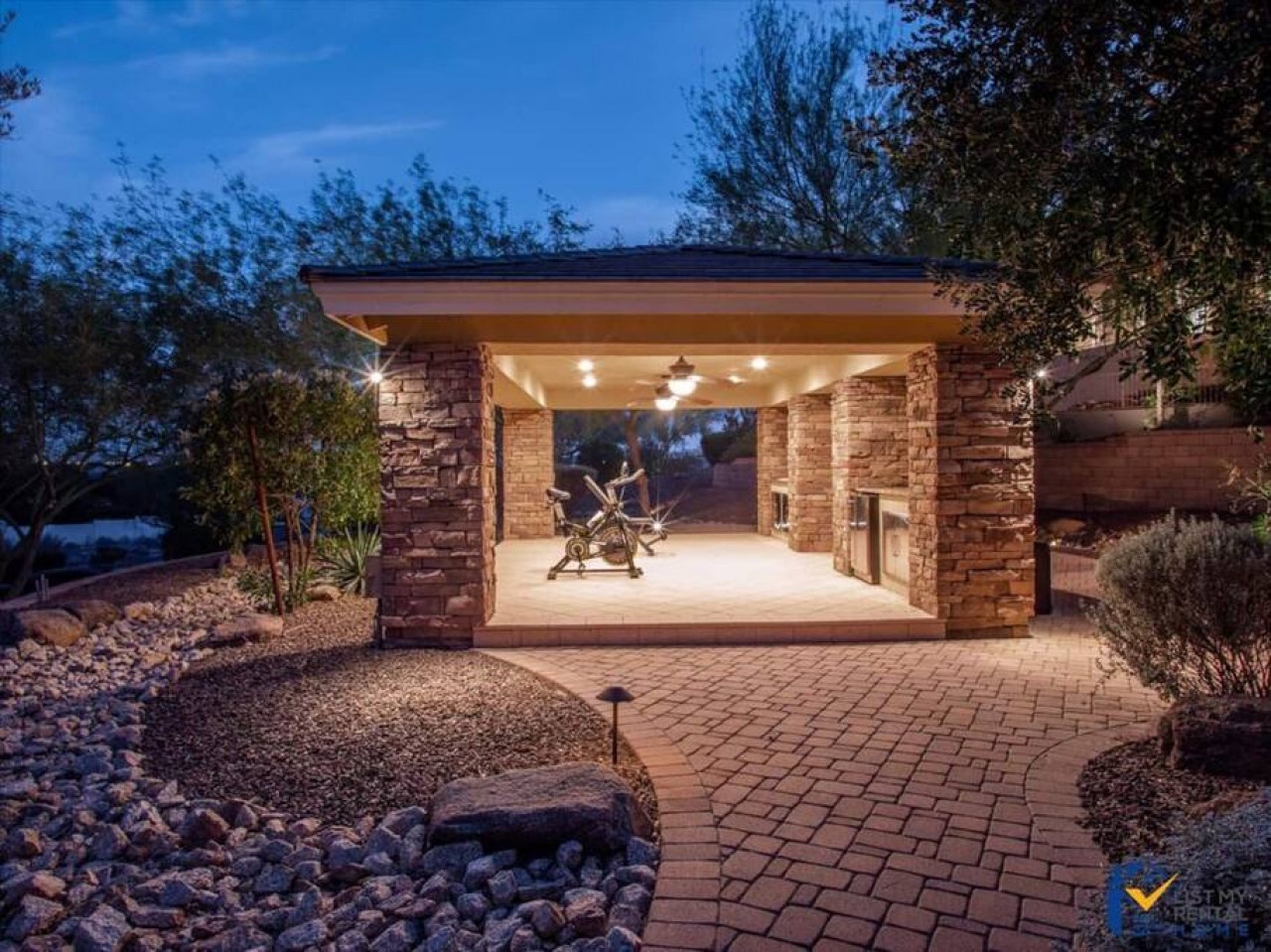 A covered outdoor fitness pavilion with stone pillars and warm lighting, set among landscaped grounds with brick pathways and desert plants, captured at twilight.