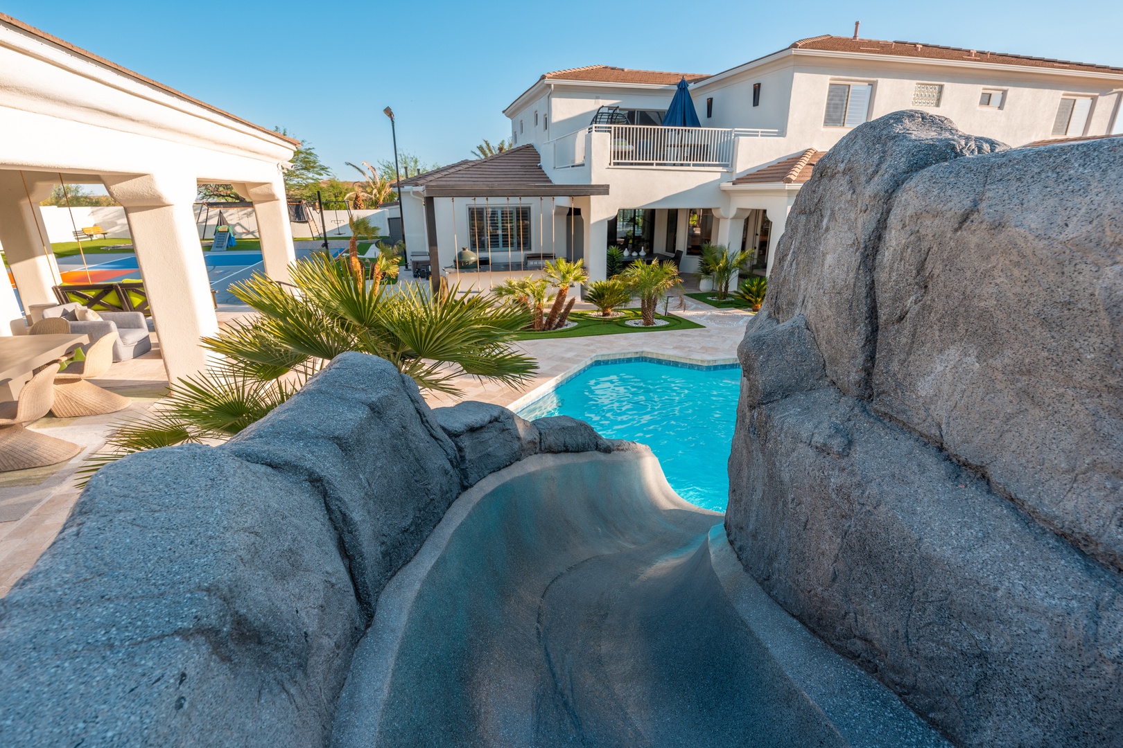 A stunning villa with a dramatic rock-feature pool, private slide, lush tropical landscaping, and covered outdoor lounge area awaits your dream vacation under sunny skies.