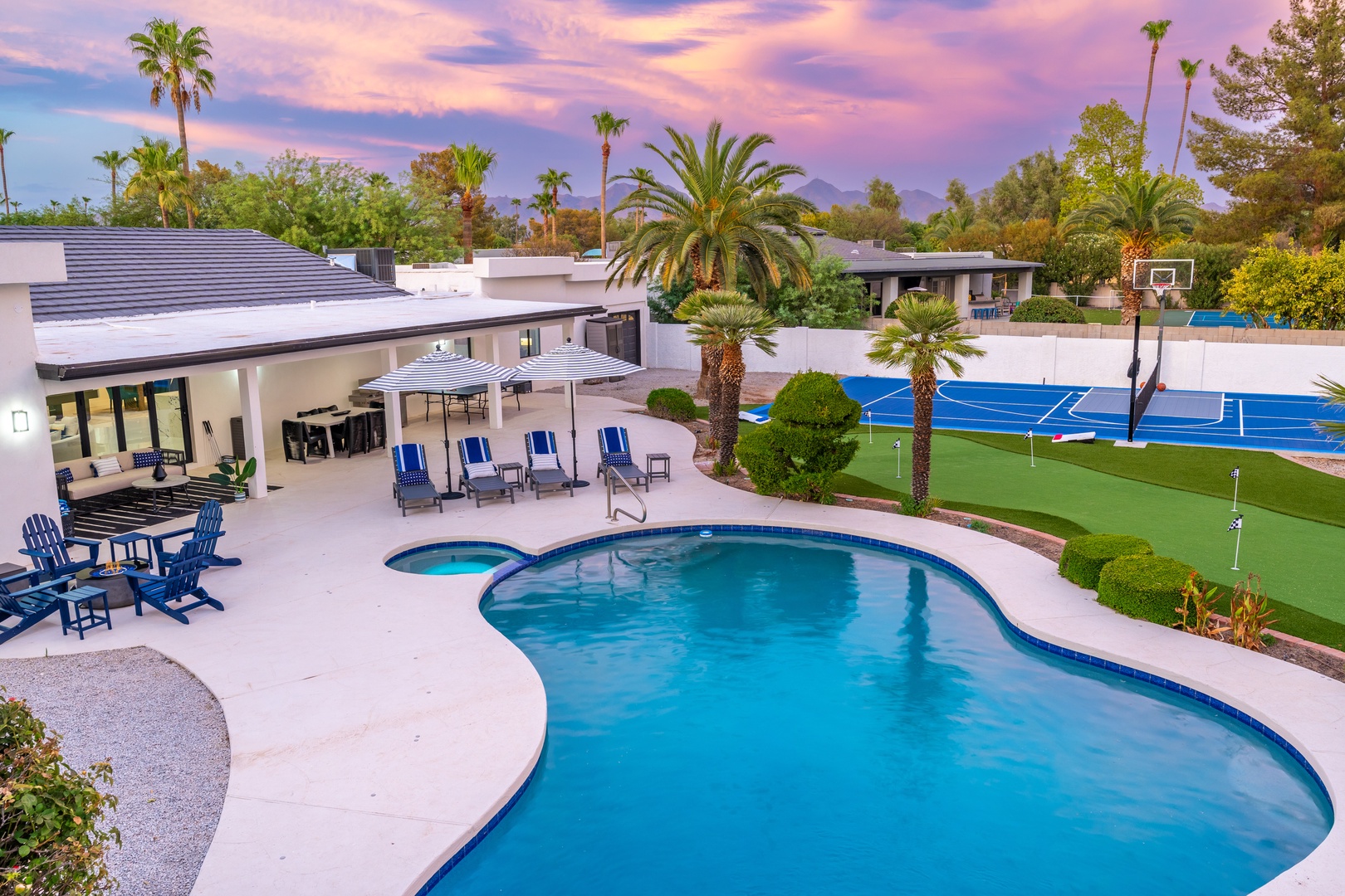 Modern desert oasis featuring a sparkling pool, hot tub, sport court, and putting green surrounded by palm trees under a breathtaking sunset—ideal for active getaways.