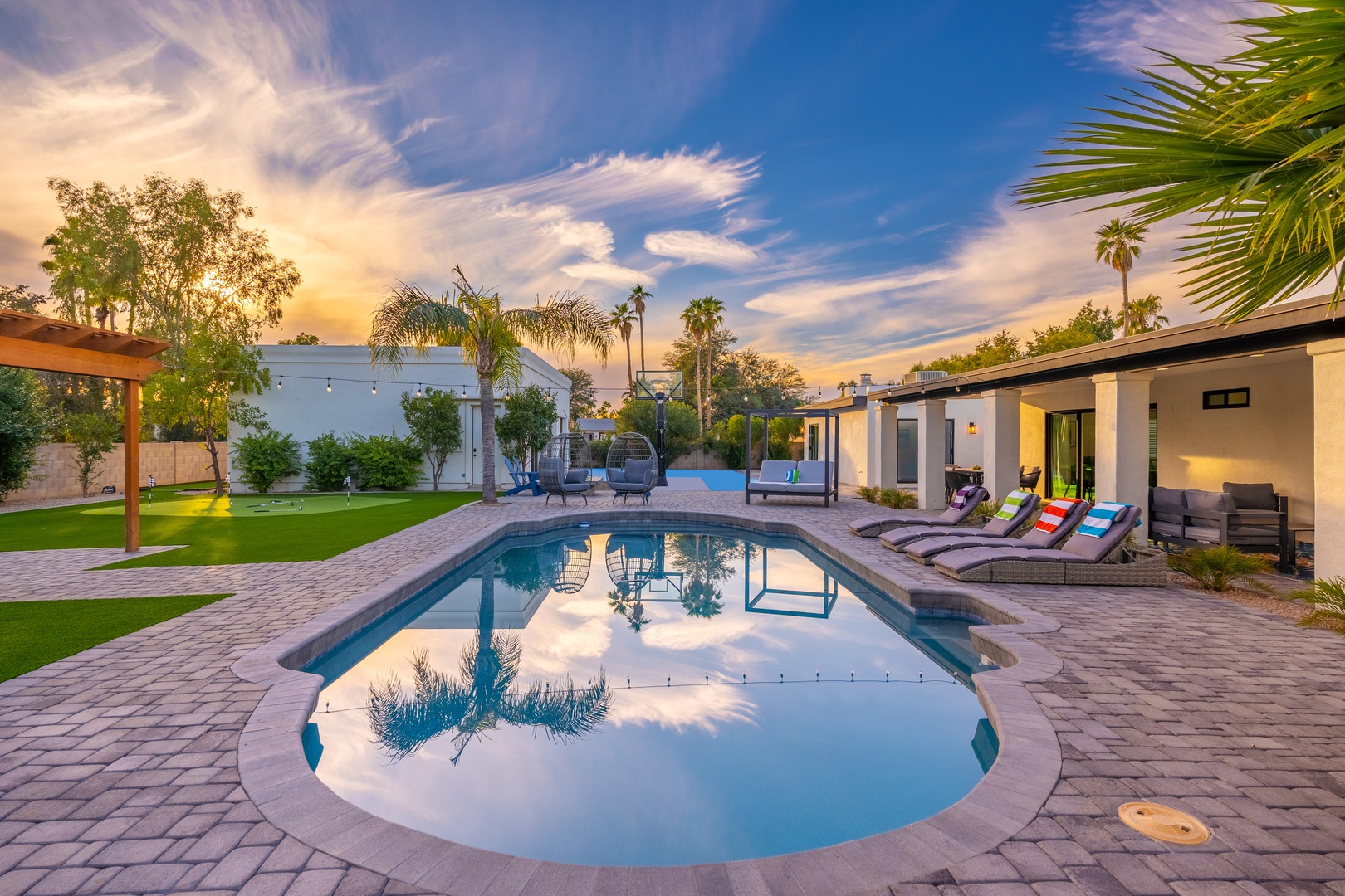 A stunning evening sky painted with warm golden hues frames this beautifully landscaped backyard retreat, where a sparkling pool reflects the surrounding palm trees and modern architecture, inviting relaxation.