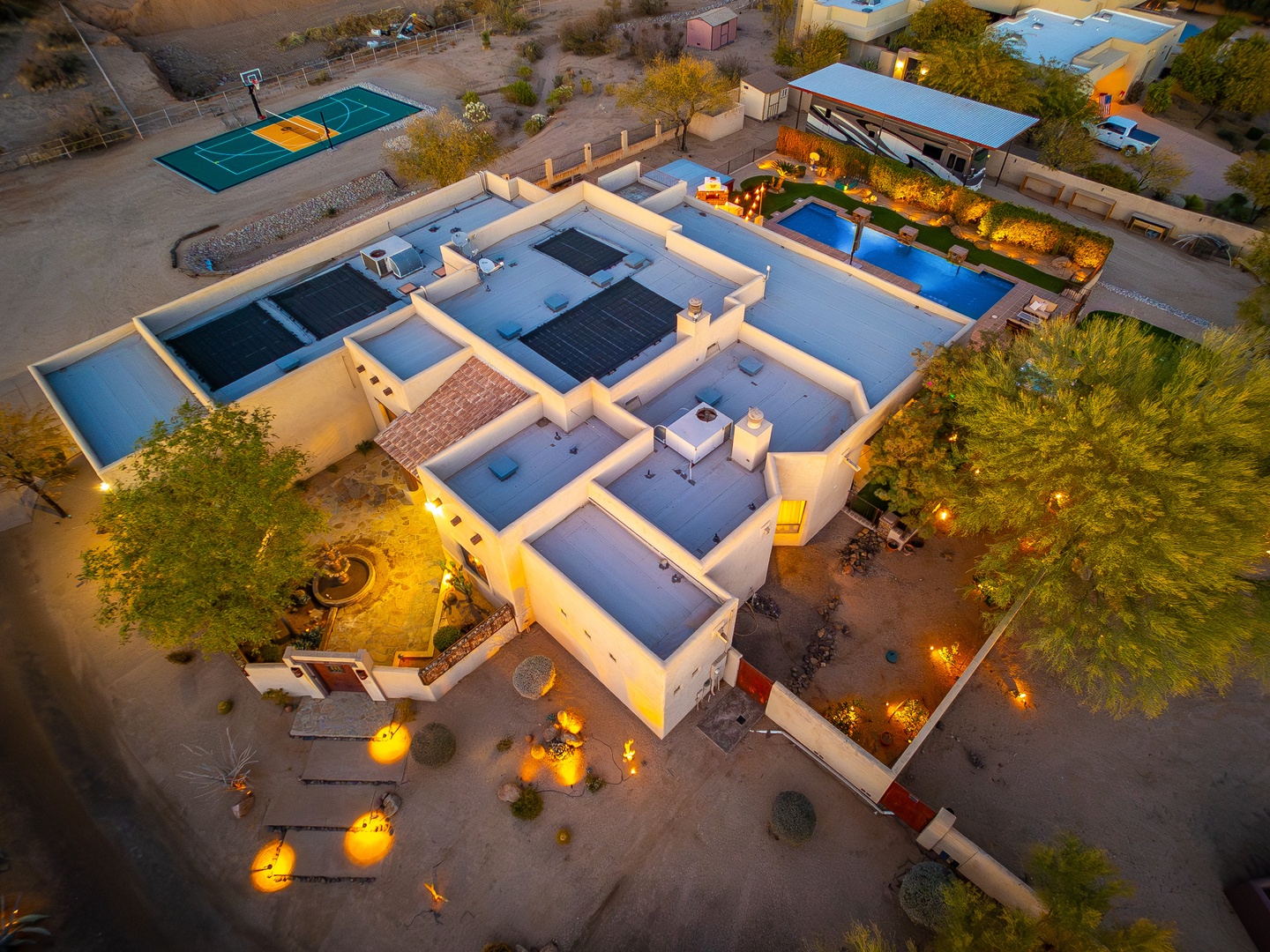 Aerial view at dusk showcasing the property's distinctive architecture, landscaped grounds, swimming pool, tennis court, and desert setting with neighboring homes.