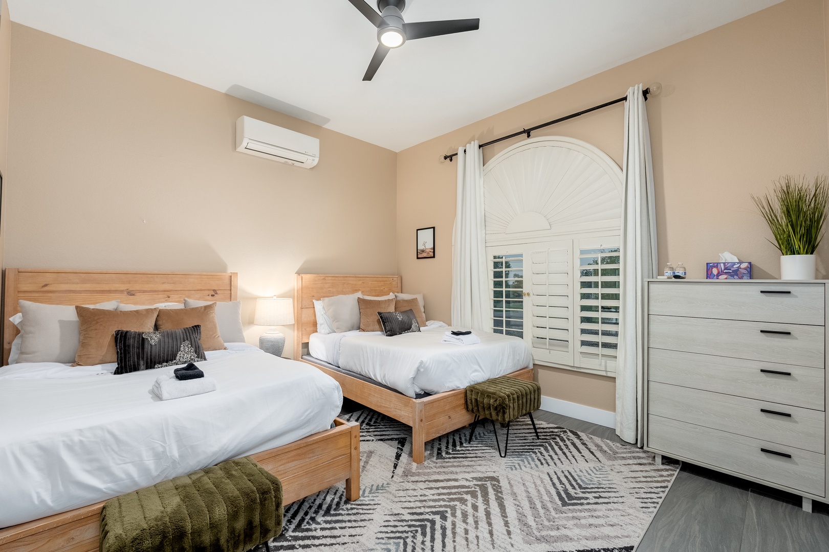 Unwind in this serene bedroom featuring two cozy beds with plush linens, modern air conditioning, and a ceiling fan to keep you cool. Natural light filters through plantation shutters, while stylish decor and ample storage ensure your comfort throughout y