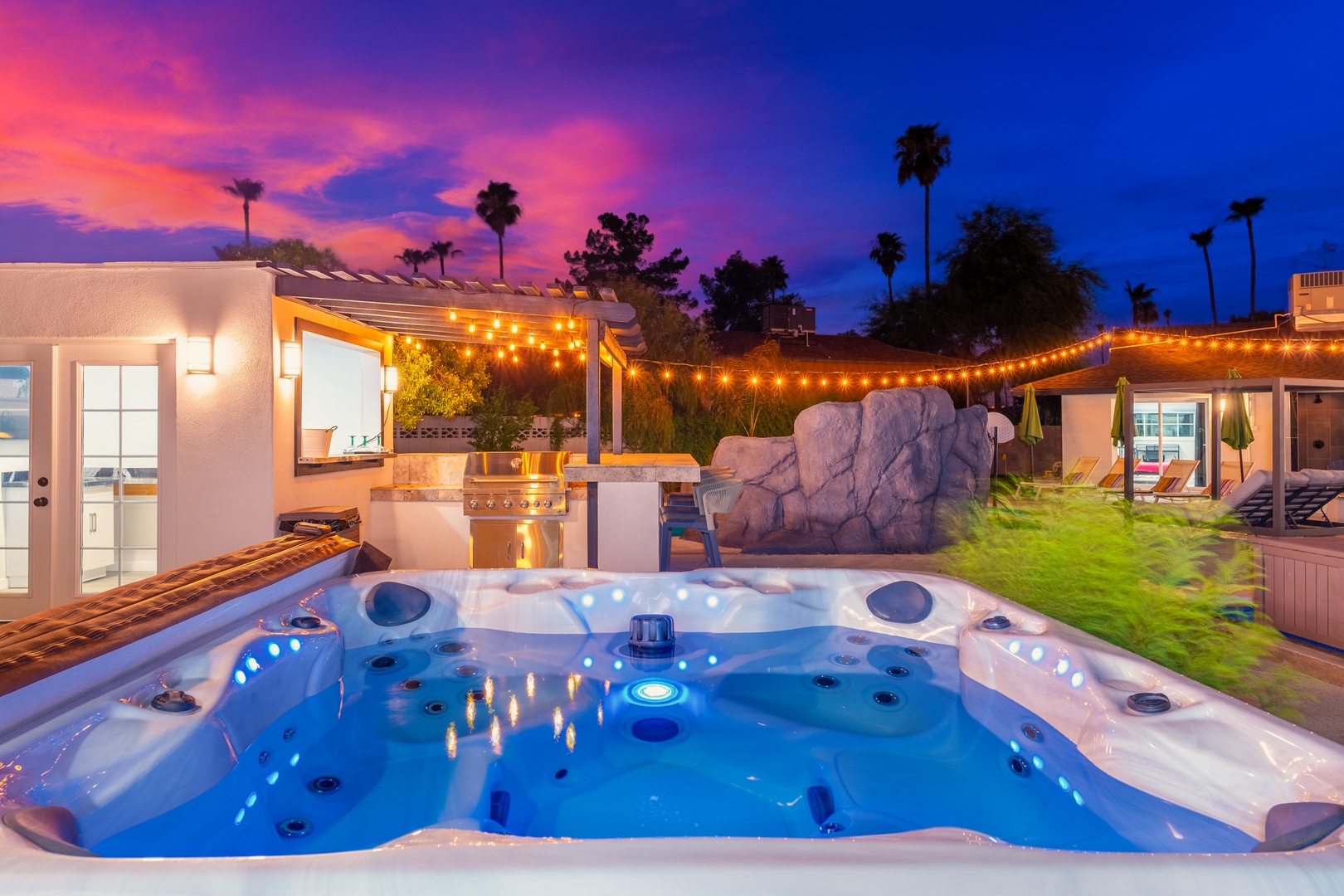 A stunning desert oasis at dusk, featuring an illuminated hot tub, outdoor kitchen, and pool area beneath vibrant pink and purple skies and swaying palms.