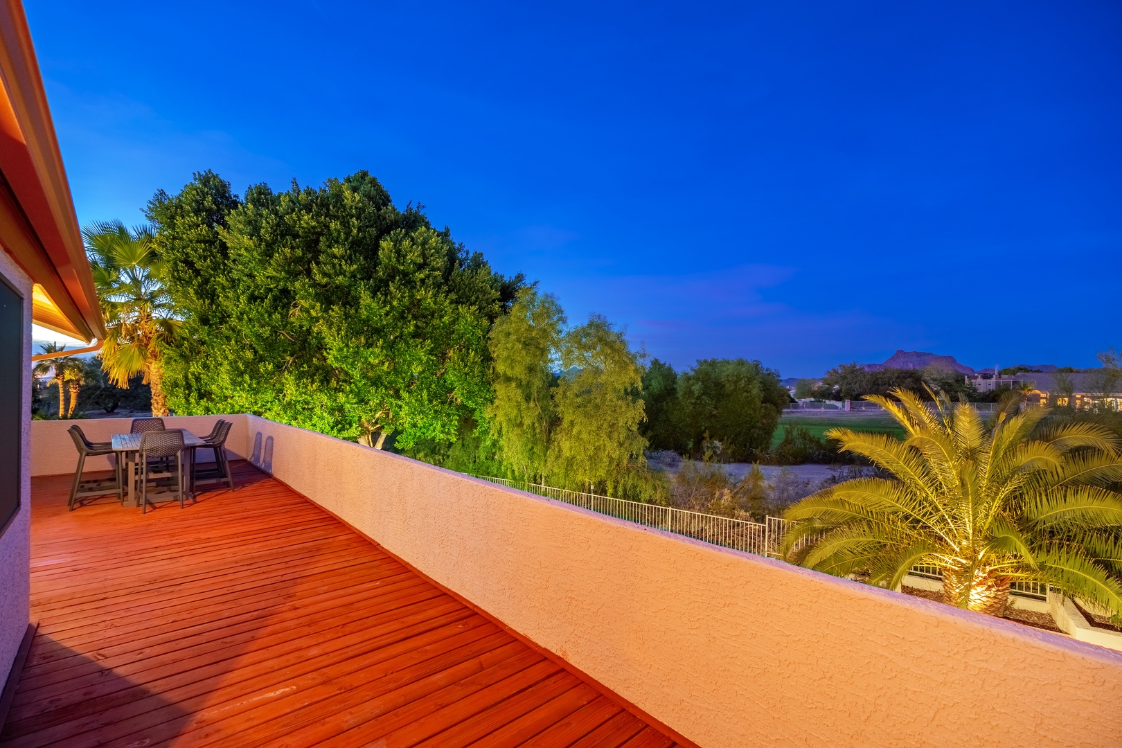 A spacious balcony with rich wood decking offers a peaceful outdoor retreat, overlooking lush greenery and mountain views. Evening lighting creates a warm, inviting ambiance for relaxed alfresco dining.