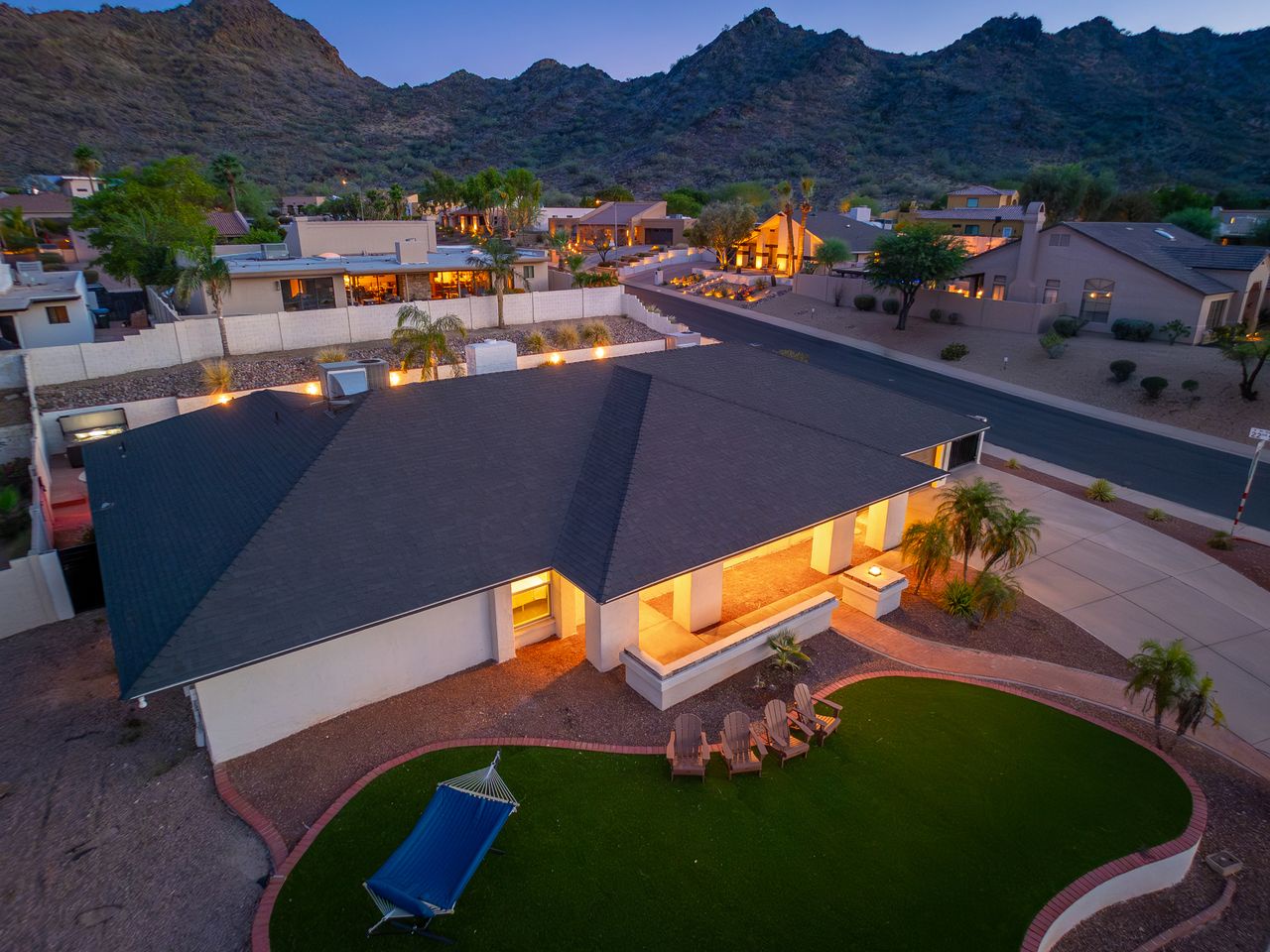 Aerial view of desert property nestled at the base of dramatic mountain peaks, with landscaped backyard and ambient evening lighting creating a warm welcome to the surrounding neighborhood.