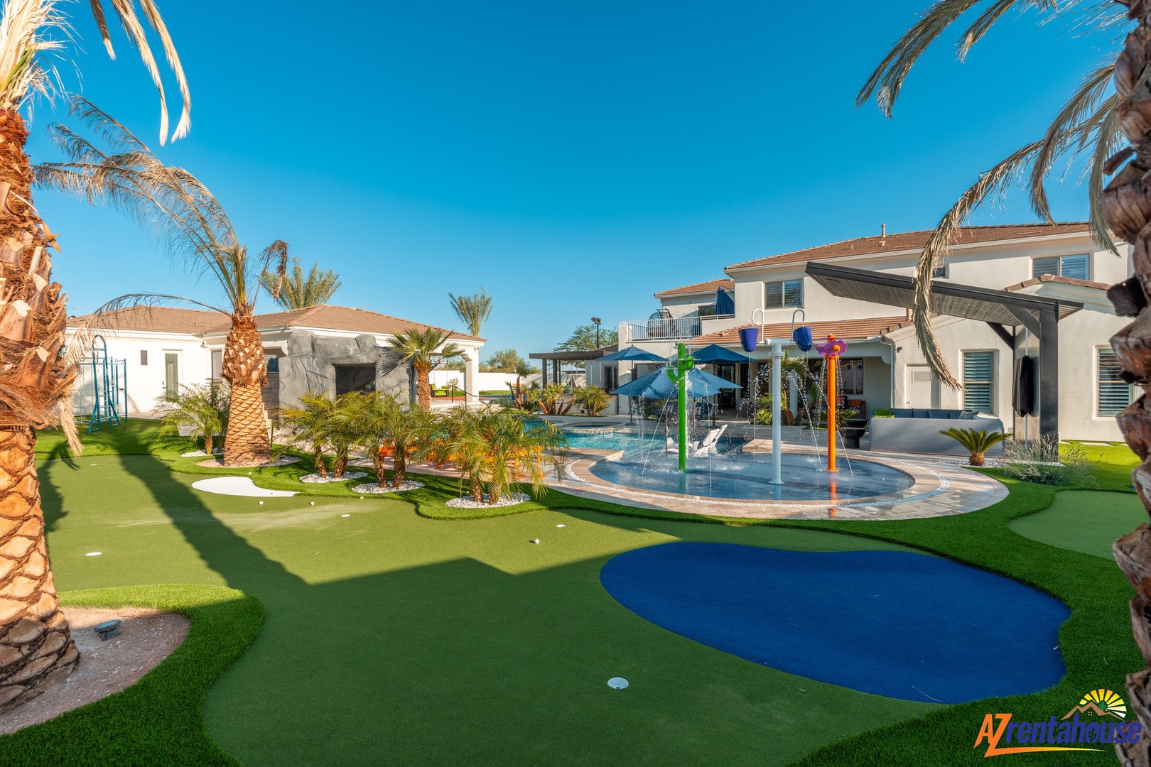 Modern villa resort featuring a splash pad, putting green, and tropical landscaping under clear desert skies.
