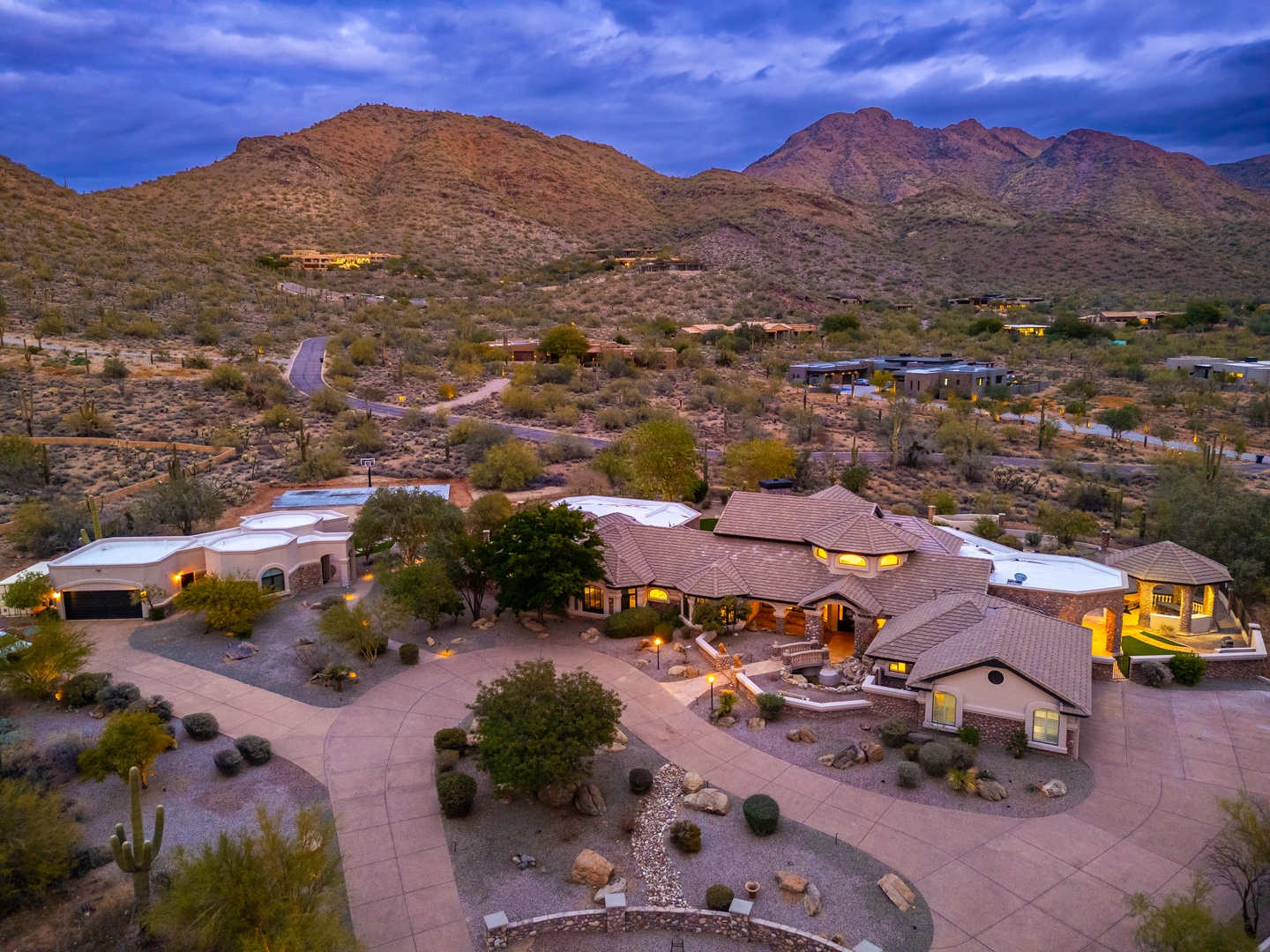An aerial view captures the property nestled against dramatic desert mountains at twilight, showcasing the estate's sprawling layout and illuminated architecture within the natural Arizona landscape.