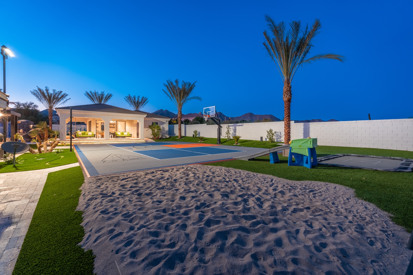 Private outdoor play zone with a beach volleyball court, full basketball court, and children's play area beneath swaying palm trees. Mountain views frame the evening scene as lights illuminate the recreational space designed for family fun.