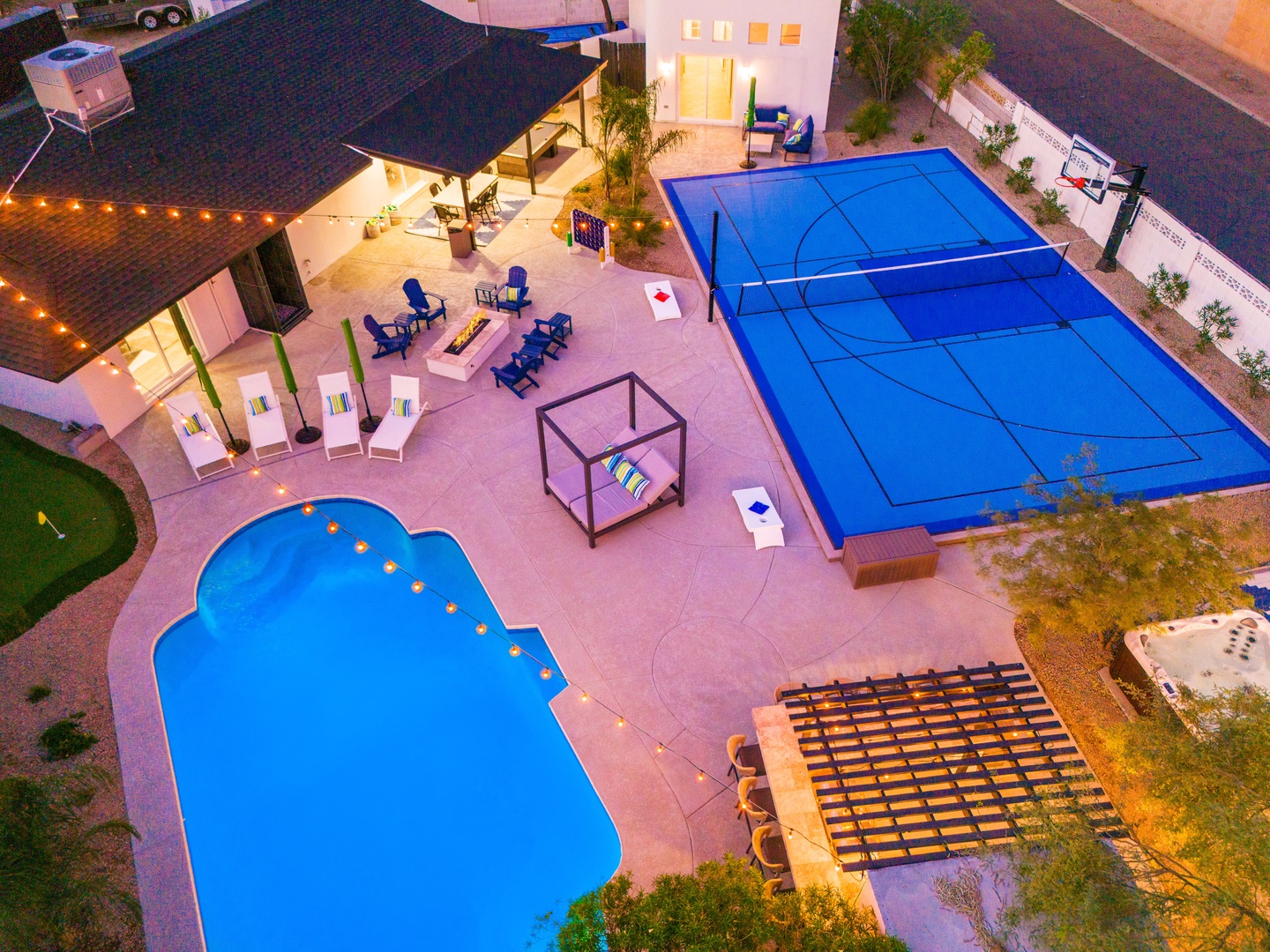 This aerial view showcases the property's extensive outdoor recreation area, featuring a private swimming pool, full-size sport court, hot tub, and multiple seating areas, set within a residential neighborhood at dusk.