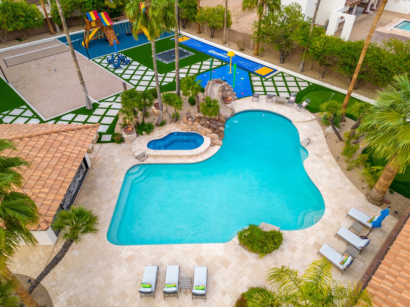 Aerial view of the property's resort-style pool area featuring a sparkling freeform pool, hot tub, colorful children's playground, and lush tropical landscaping with palm trees and lounge seating throughout.