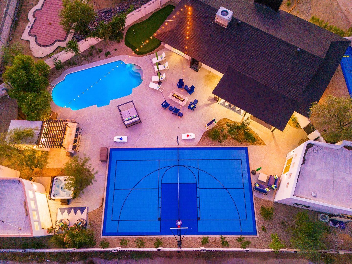Aerial view of the property showcasing a full outdoor basketball court, swimming pool, hot tub, covered patio, and manicured grounds illuminated at dusk.