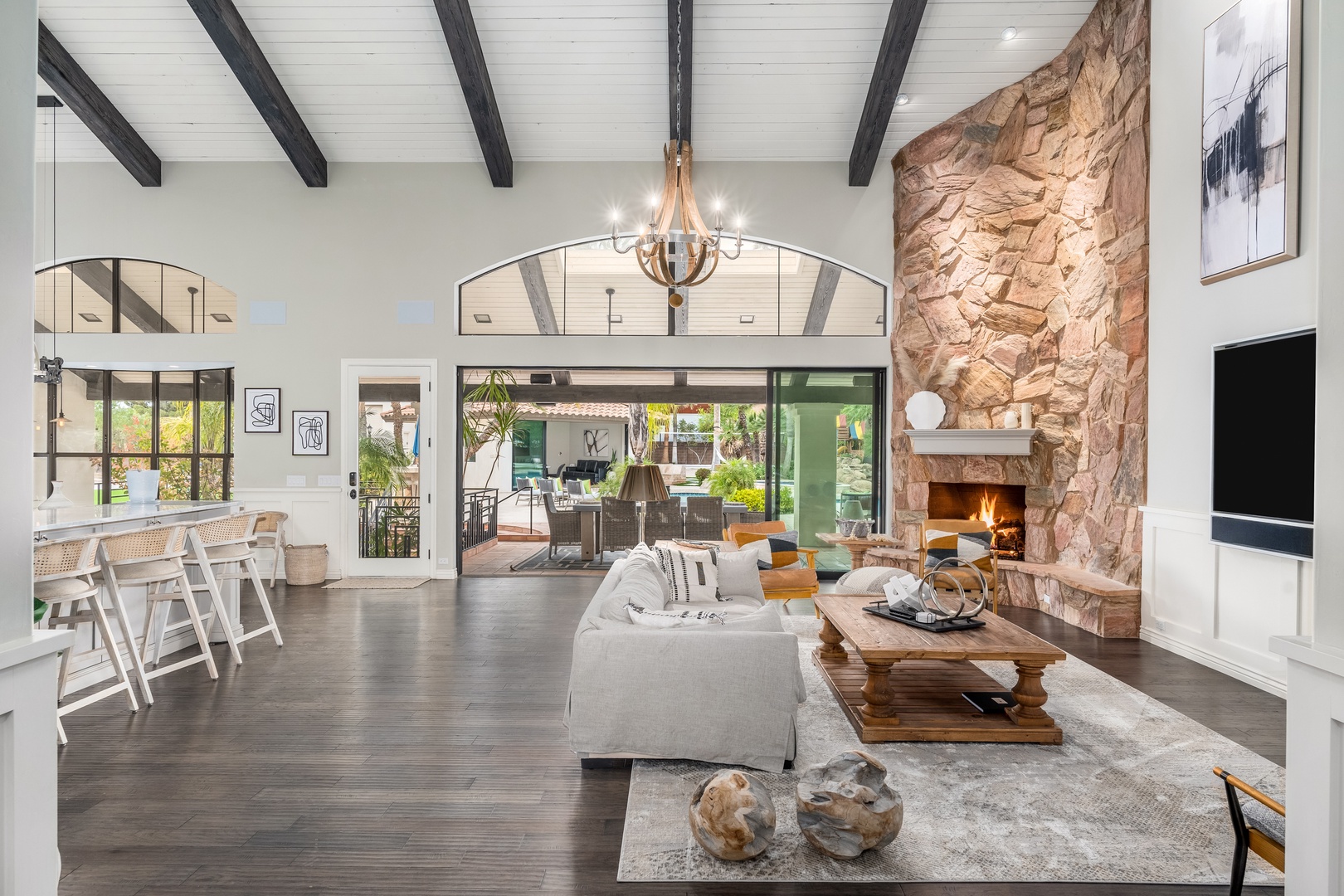 Relax in this stunning open-concept space featuring soaring ceilings with exposed beams, a majestic stone fireplace, and seamless indoor-outdoor flow through expansive glass doors opening to your private patio.